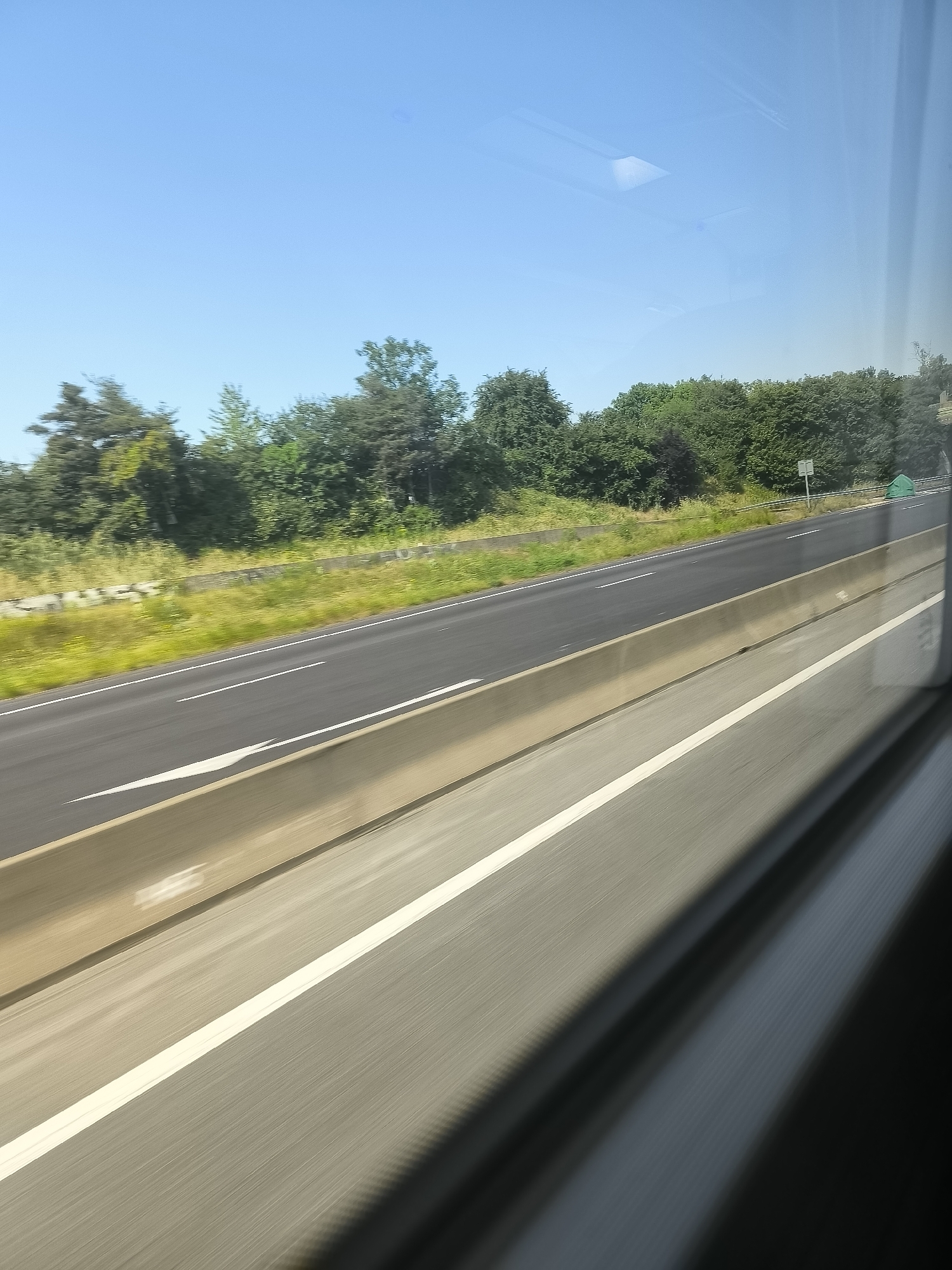 The image depicts a view from a moving vehicle, likely a car or train, traveling on a highway near L'Horme, France. The highway is visible, with clear lane markings and a concrete median barrier separating the two directions of traffic. Beyond the highway is a grassy verge, followed by a dense line of trees and bushes, typical of the French countryside. The sky is clear and blue, suggesting a sunny day. Reflections can be seen in the window of the vehicle, indicating the image was taken from inside. The primary focus is on the road and surrounding environment, with no individuals immediately visible in the frame.