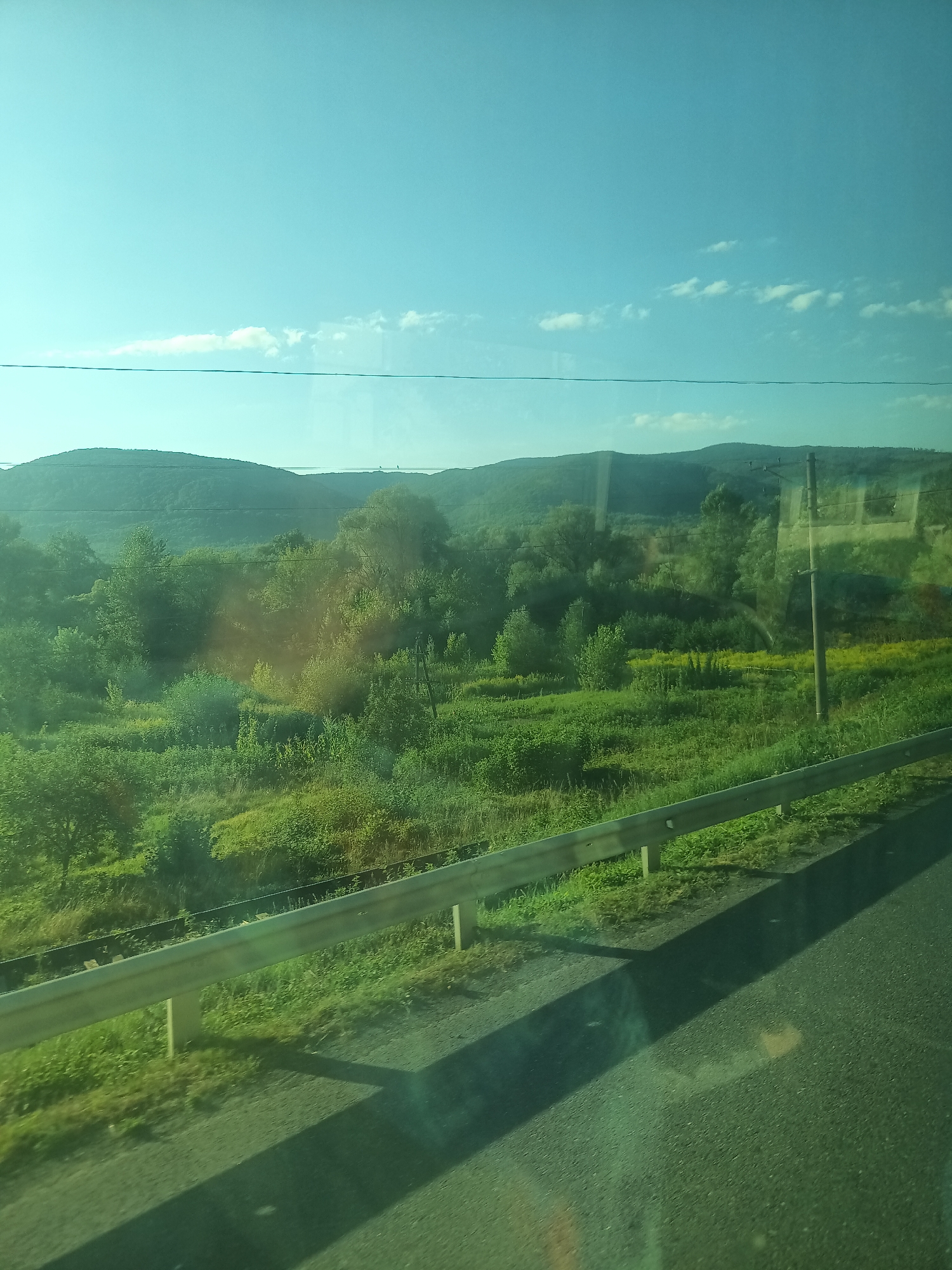 The image captures a view of rolling green hills and a clear blue sky with sparse clouds. A metal guardrail runs along the edge of a paved road in the foreground, casting a long shadow. Beyond the guardrail, a lush, verdant landscape stretches out, dotted with trees and patches of yellow wildflowers. In the distance, the hills are covered in dense foliage, and a single utility pole stands on the right side of the frame. The scene appears to be a roadside view of a rural area, possibly on the outskirts of Perechyn, Ukraine, given the geographical context.