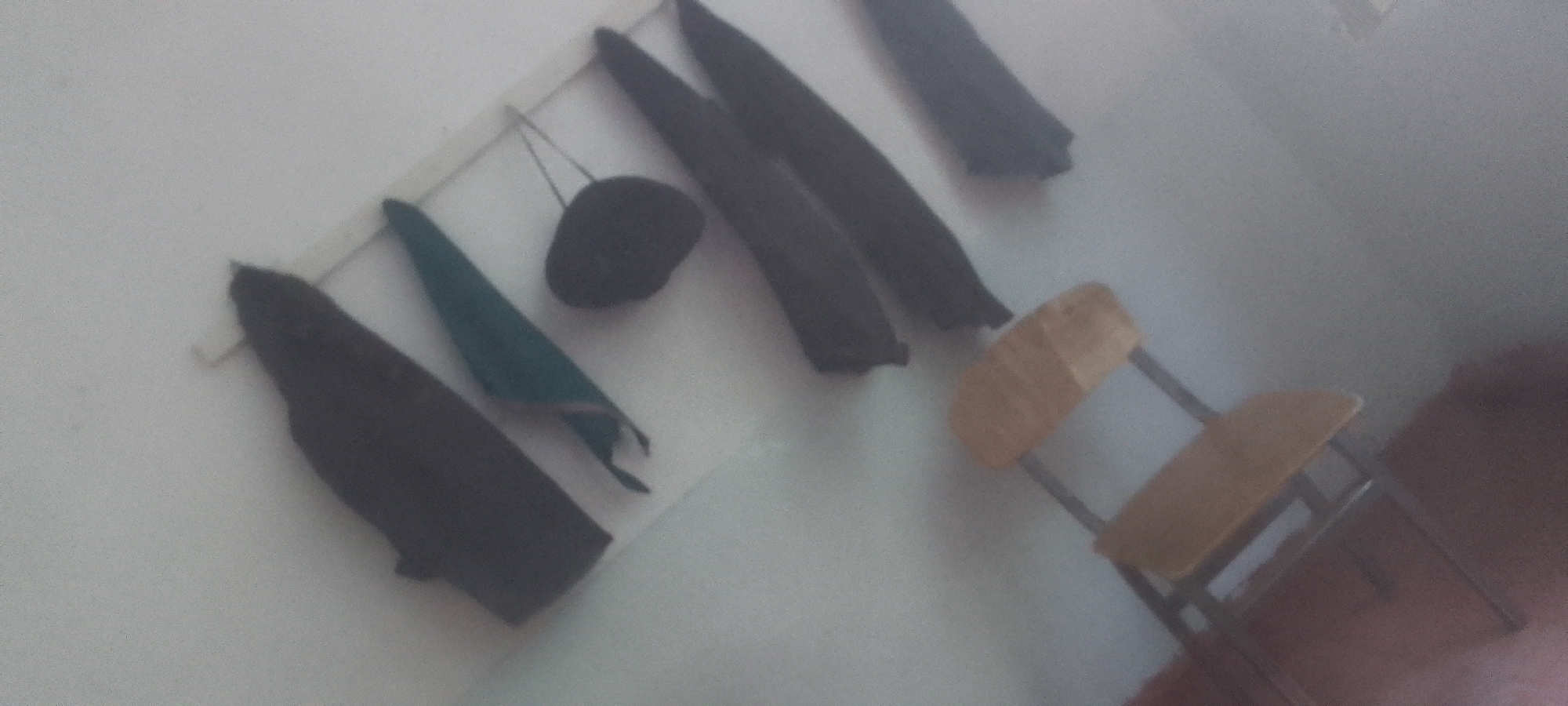The image shows a wall with several dark-colored garments hanging on it. One garment is a dark blue or black coat, long and full, hanging on a hook. Next to it, there is a smaller dark object, possibly a hat, suspended by a string. To the right of that, there are more dark garments, one appearing to be a dark green scarf or fabric. Further to the right, there are two more long, dark garments hanging. In the foreground and to the right, a light brown wooden chair with a metal frame is visible. The background is a light-colored wall. The image appears to have been taken in an indoor setting, possibly a room in Dushanbe, Tajikistan, given the context of the request. The lighting is dim, and the image quality is somewhat grainy.