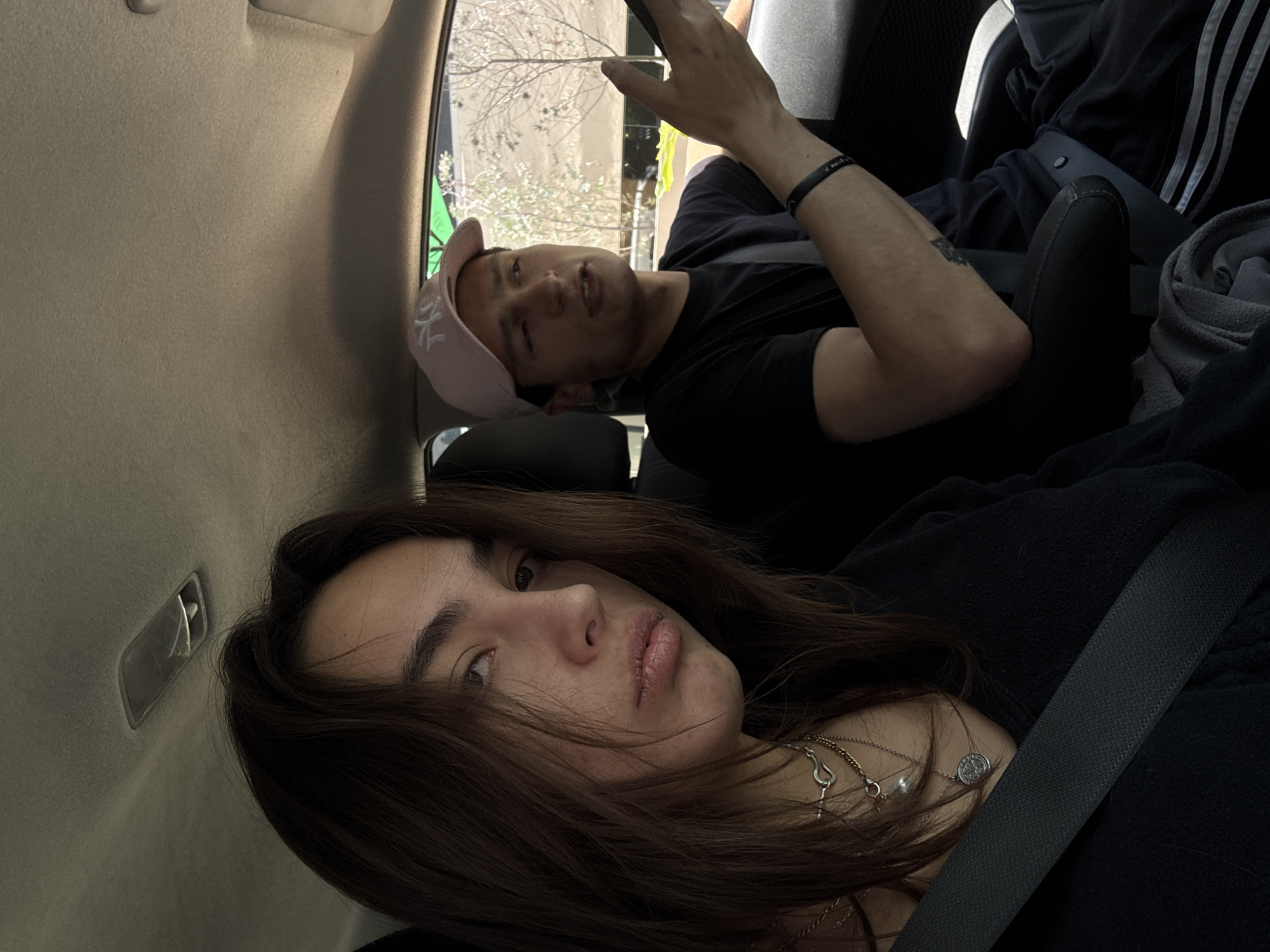 Two individuals are inside a vehicle, wearing their seatbelts. The person in the foreground has long brown hair and is wearing multiple silver necklaces. The individual in the background is wearing a pink baseball cap, a black t-shirt, and a black wristband. Through the car window, trees and a building are visible. The location is suggested to be Mexico City, Mexico.