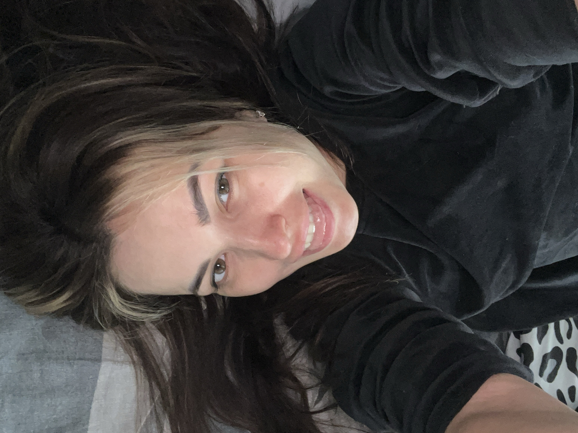 A woman is lying down, looking up and smiling at the camera. She has brown hair with blonde highlights near her face, light skin, and brown eyes. She is wearing a dark-colored, possibly velvet, top. She is lying on a gray and white patterned surface, possibly a bed. The location is Unknown, Unknown.