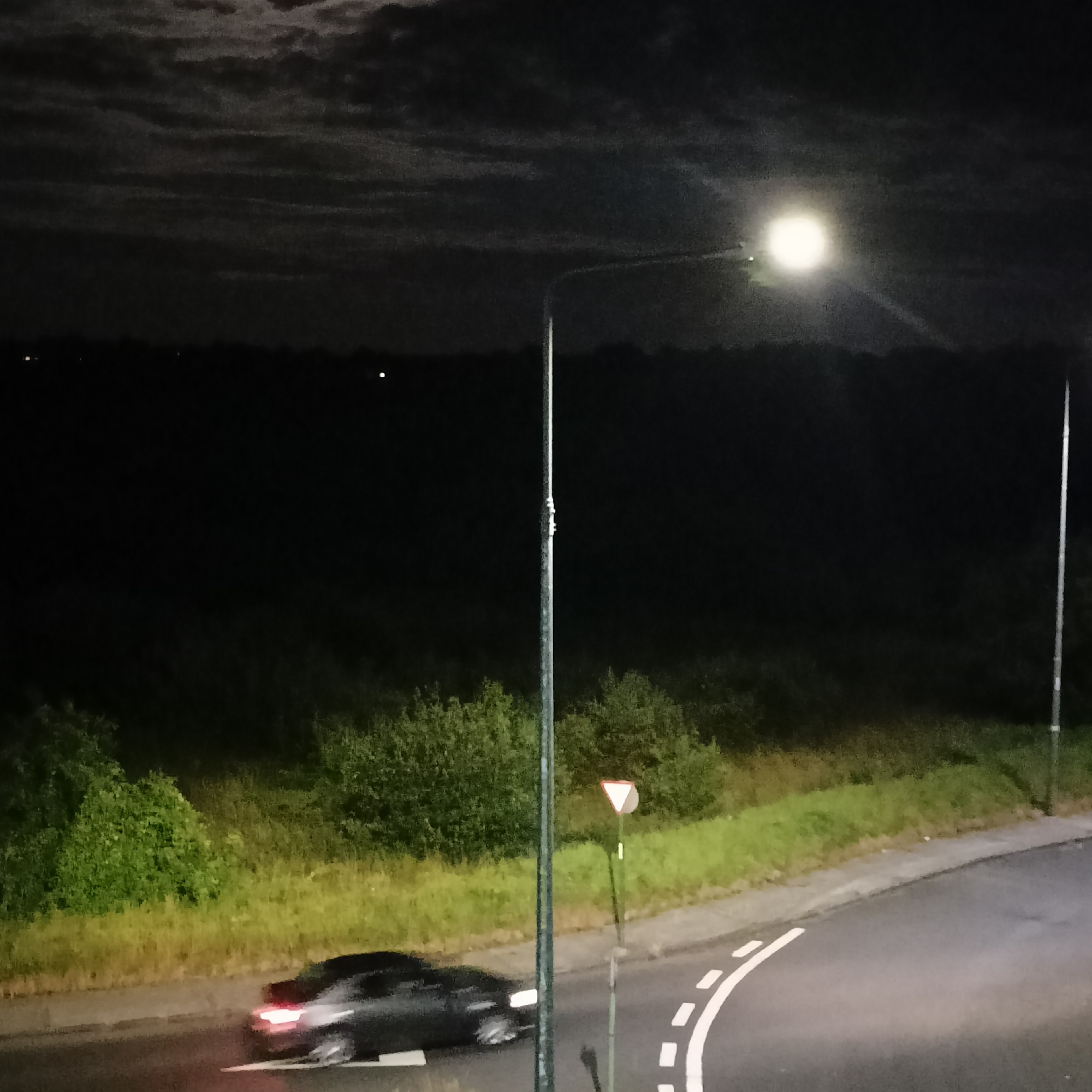 The image depicts a night scene on a road, likely in a rural or suburban area near Limerick, Ireland. A dark-colored car is visible driving on the asphalt road, with its headlights illuminated and rear brake lights glowing red. The car appears to be in motion, suggested by a slight blur. To the right of the road, there is a triangular yield sign mounted on a pole, and a streetlamp with a bright light source is positioned in the center of the frame. The road curves to the right, bordered by a grassy verge and lush green foliage. In the background, a dark expanse of trees and fields stretches towards the horizon. The sky is cloudy, with the moon or a bright celestial body partially obscured by the clouds, casting a subtle glow. There are no visible people in the image. The overall atmosphere is dim, illuminated primarily by the streetlamp and the car's lights.