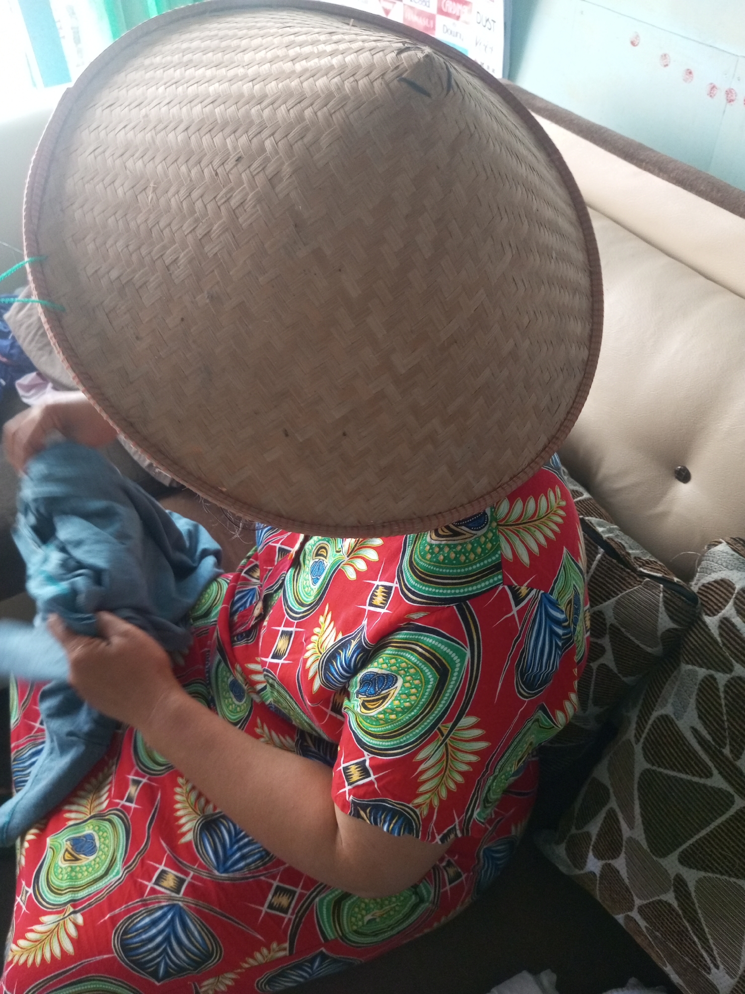 In Andongsili, Indonesia, a person wearing a woven conical hat is seated indoors. The hat obscures their face and is made of a light brown, woven material. The person is wearing a vibrantly colored shirt with a red base and a pattern of green, blue, and white shapes that resemble eyes or leaves. They are holding a crumpled piece of light blue fabric in their hands, appearing to be folding or manipulating it. The background shows a beige sofa with dark patterned cushions, suggesting a domestic setting. A wall with some faint markings is visible in the upper left corner. The lighting indicates an indoor environment.