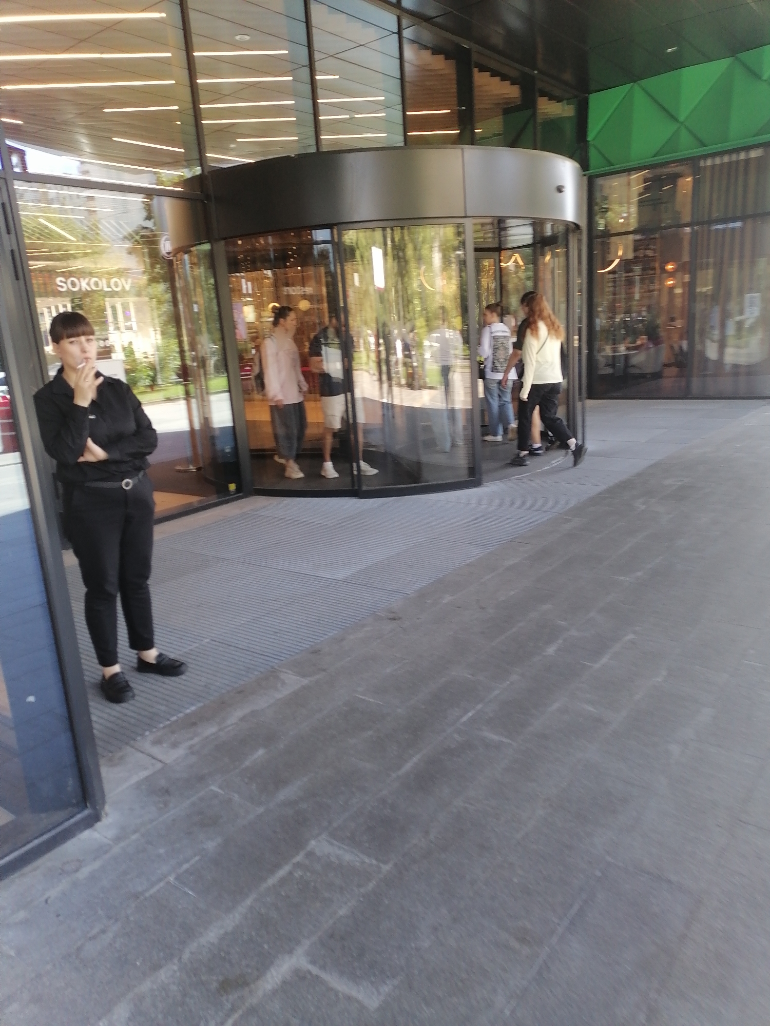 A woman stands in front of a revolving door entrance to a building, smoking a cigarette. She is wearing black pants and a black shirt, with her arms crossed. Inside the revolving door, several people are visible, including a woman in a pink shirt and a man in a white shirt and shorts. The building appears to be a modern commercial establishment, possibly a retail store or office building, with a glass facade and a green geometric design element on the exterior. The signage "SOKOLOV" is visible on the building. The scene is set outdoors on a paved area, suggesting a public space. The overall impression is one of a typical urban environment. The image was taken in Moscow, Russia.