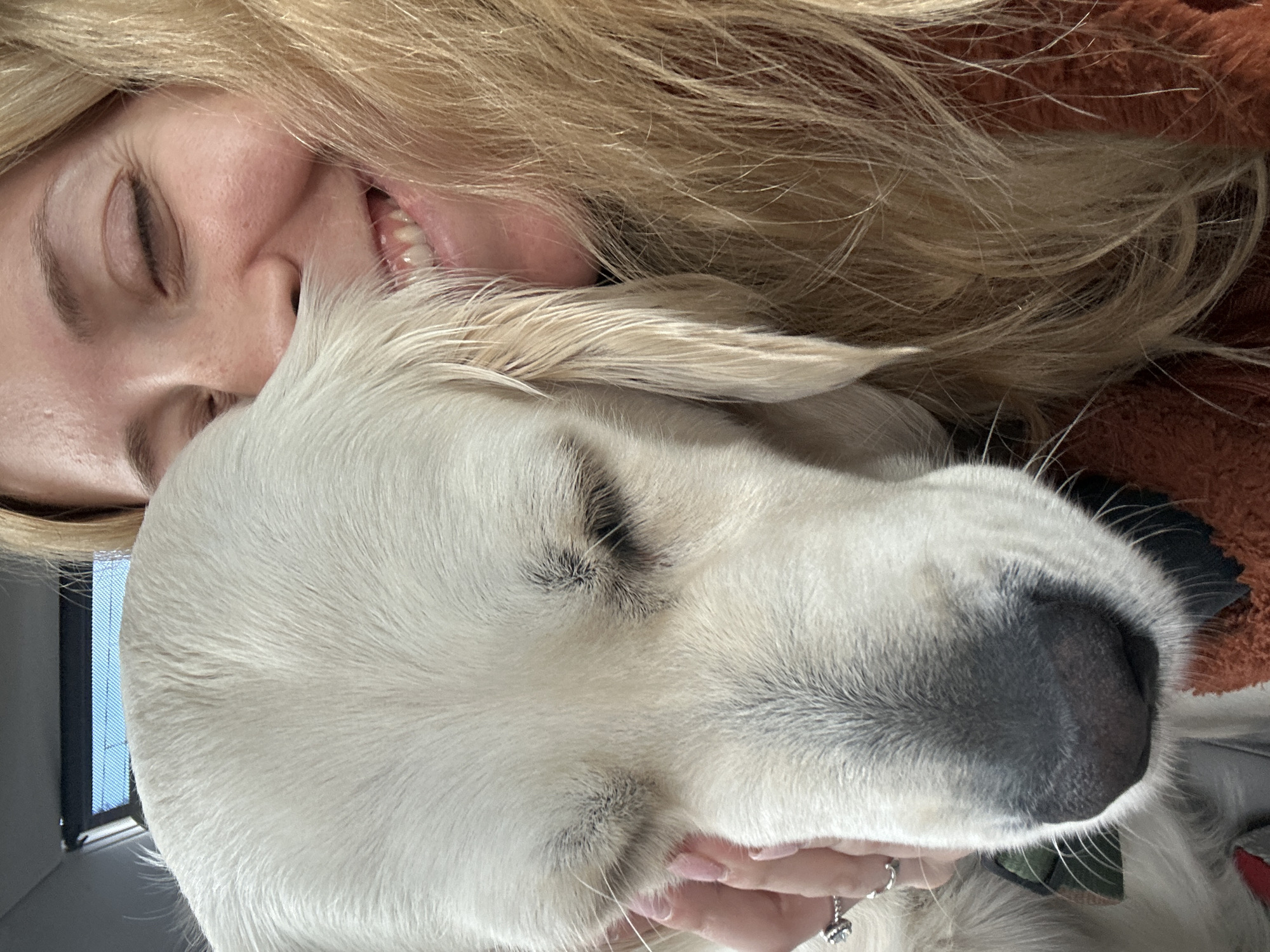 A woman with blonde hair and closed eyes is smiling while embracing a light-colored dog with its eyes closed. The dog's nose is prominent and dark. The woman's hand is visible, wearing a ring. A window frame is partially visible in the background. The location cannot be determined from the image.