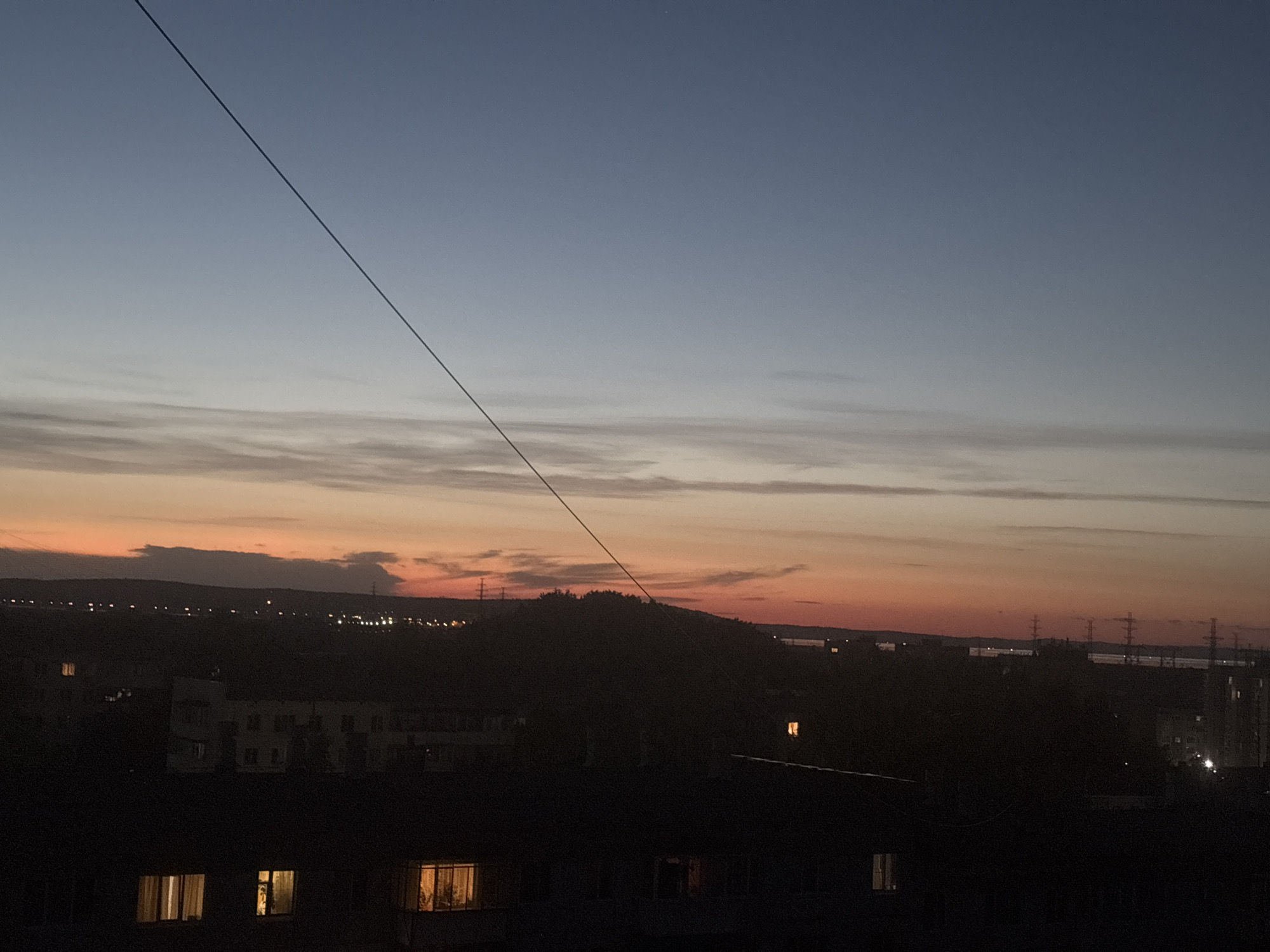The image depicts a cityscape at dusk. The sky transitions from a gradient of blue to orange near the horizon. A dark, thin power line stretches diagonally across the frame. Below the horizon, there's a silhouette of a landmass, possibly a hill or a line of trees. A row of buildings is visible, with illuminated windows suggesting that it's late in the day. The overall scene gives the impression of a quiet urban setting at twilight.