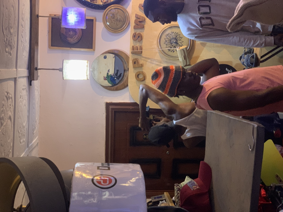 The image shows the interior of a shop, with the address being Ita Oniyan, Nigeria (latitude: 7.282, longitude: 5.161). There are three men in the shop, one of whom is wearing a white tank top and is facing away from the camera. The other two men are facing the camera, and one is wearing a pink tank top and a multi-colored beanie hat. The man in the beanie is touching his hat with his right hand. The other man is wearing a gray sweatshirt with the word "DOU" on it. The shop is decorated with wooden wall art, including a circular framed picture with a man in it, a sunflower-shaped piece of art, and the word "OPESWA" in wooden letters. The shop also has two pendant lights, one of which is on, and a number of hats and other objects sitting on a shelf. There is a closed wooden door to the left side of the image. There are sandals on a shelf in the foreground of the picture. There is a large, open ceiling fan in the corner of the image, with a gray metal covering around it. The image is lit by overhead lights and the daylight that is coming through the doorway in the background.