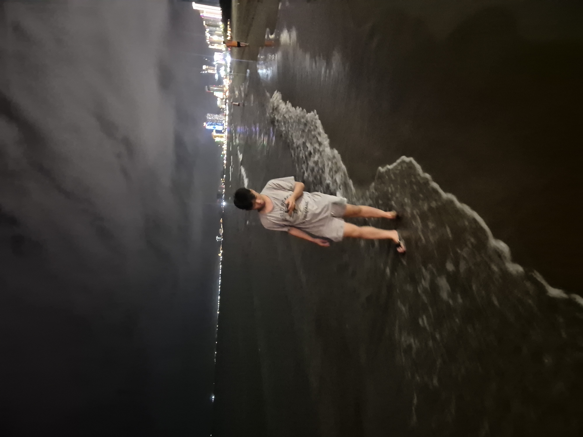 This is a nighttime photograph of a person walking on a beach in Đà Nẵng, Vietnam, near the city skyline. The person, appearing to be a young male, is wearing a grey t-shirt and grey shorts. He is wading into the shallow water at the edge of the ocean, with small waves lapping at his feet. He is looking down at his phone. In the background, the illuminated skyline of Đà Nẵng is visible across the water, with tall buildings and lights. The sky is dark and cloudy. There are other indistinct figures visible further down the beach. The sand is dark and wet, and the ocean appears calm.