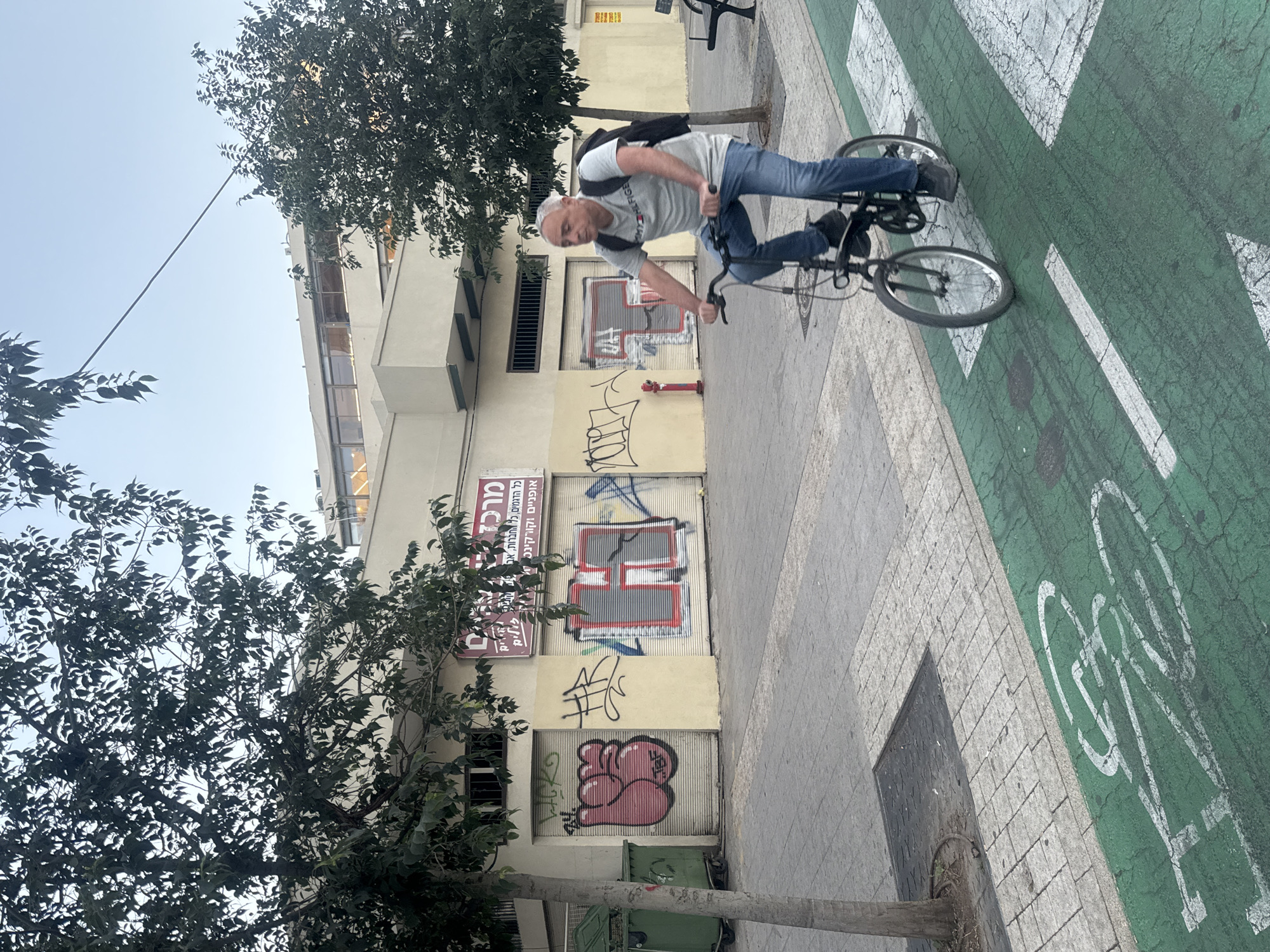 This image depicts an urban street scene in Tel-Aviv, Israel, captured during the daytime under a clear sky. The photo is rotated 90 degrees clockwise, presenting the street and building vertically.
The central subject is a man riding a bicycle. He appears to be middle-aged, with short, light-colored hair and stubble, wearing a light gray t-shirt with a partial "TOMMY HILFIGER" logo, blue jeans, and a dark backpack. He is leaning forward on a dark-colored bicycle, actively pedaling.
He is positioned on a green-painted bike lane, clearly marked with white arrows and a white bicycle symbol. Adjacent to the bike lane is a gray paved sidewalk.
In the background, a light-colored, multi-story building lines the street. Its lower level features several roll-down shutters, some heavily covered in graffiti, including a red and gray geometric pattern and a pink, bulbous graphic. A red fire hydrant is visible on the building's wall. A sign above the shutters displays Hebrew text, partially decipherable as "מרכז" (Center) and mentioning "אופניים" (bicycles) and "היבוא" (import).
Green foliage from several trees frames the scene, with one large tree prominent on the left and another partially visible on the right, casting shadows and adding to the urban landscape. The sky is clear blue, indicating fair weather. The man's expression is neutral, focused on his activity.