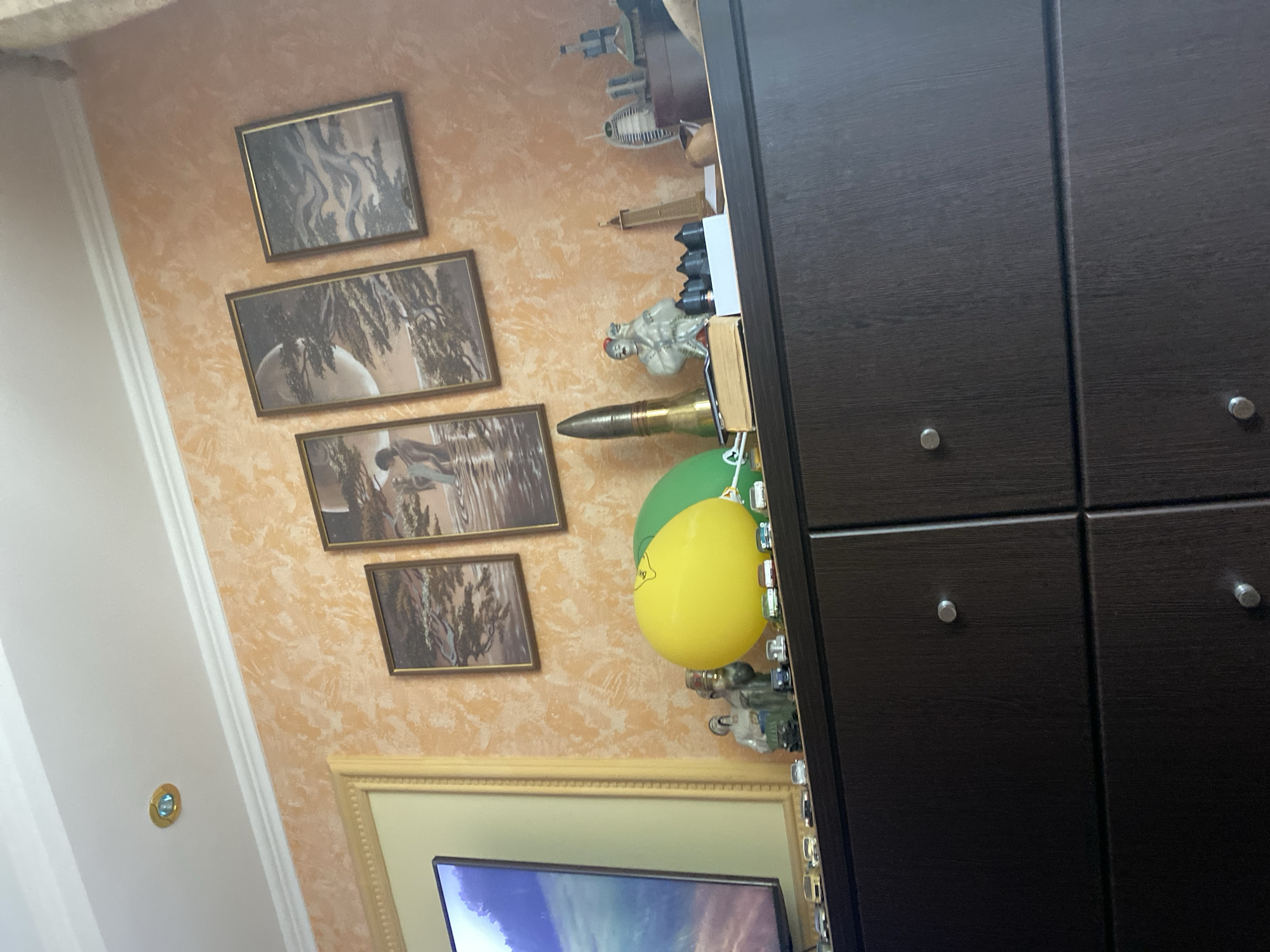 The image shows a dark brown dresser with a collection of items on top. The items include several framed paintings on an orange textured wall, a large yellow balloon, a green balloon, a model of a projectile, a figurine of a man, and several small model cars. There is also a television screen on the left side of the frame, showing a landscape scene. In the background, part of the ceiling with a light fixture is visible. The overall impression is of a home interior.