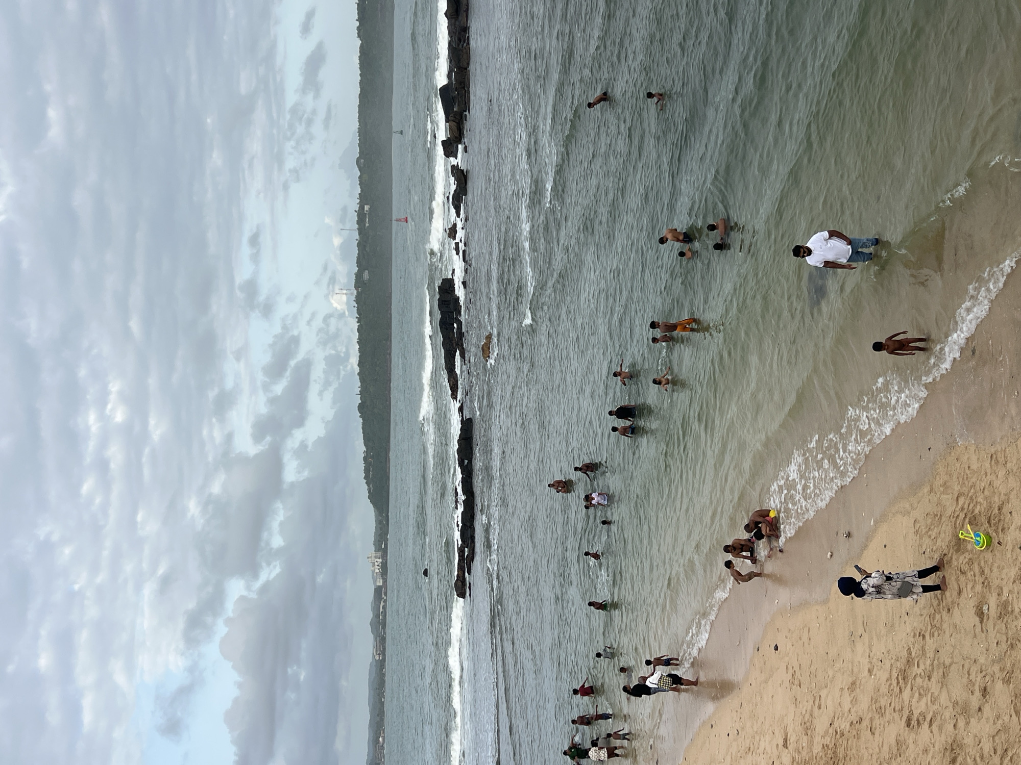 The image depicts a beach scene in Galle, Sri Lanka, under a cloudy sky. People are swimming and wading in the water near a rocky outcrop that extends into the sea. Closer to the shore, there are more people on the sandy beach, including a person lying down. A yellow and green bucket sits on the sand. The water appears somewhat murky, and there are gentle waves breaking along the shoreline. In the background, there is a coastline with buildings and vegetation. A red navigational marker is visible in the water near the rocky outcrop.