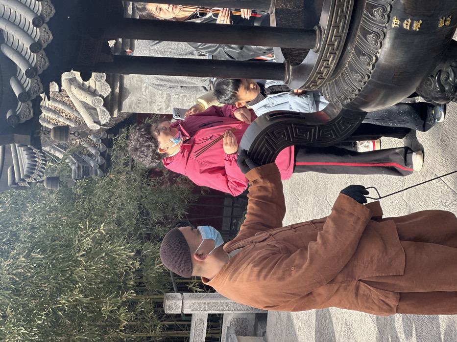The image, rotated 90 degrees counter-clockwise, depicts an outdoor scene at what appears to be a temple or traditional cultural site, with several individuals participating in an activity involving incense. The setting is bright and sunny.
In the foreground, on the right, a person is seen in profile, wearing a brown, traditional-style garment resembling a monk's robe, a dark brown knitted cap, and black gloves. A light blue surgical mask is pulled down below their chin. Their left hand rests on a large, dark, ornate structure, which features a distinctive geometric meander pattern. This person is holding a black cord or string in their right hand, which extends downwards out of frame. Behind them, a stone railing with carved details is visible.
In the mid-ground, slightly to the left of the center, a woman with short, curly dark hair is prominent. She is wearing a bright pink jacket, black pants with a red stripe down the side, and a light blue surgical mask covering her mouth and nose. She holds several sticks of lit incense in her right hand, positioned near her chest, suggesting an act of offering or prayer. Her left hand holds a smartphone, which rests on her hip.
Behind the woman in pink, a young boy with dark hair is partially visible, wearing a light blue jacket. Further in the background, another woman is seen behind a dark horizontal bar, also holding sticks of incense. She has dark hair and wears sunglasses pushed up onto her head.
The large, ornate structure in the mid-ground appears to be made of dark metal or stone and features elaborate carvings, including the meander pattern. On its upper part, faint gold-colored Chinese characters are visible, which appear to read "白甘" (bái gān), possibly part of a larger inscription.
The background on the left features lush green bamboo foliage, suggesting a garden or natural area within the site. Above the bamboo, traditional East Asian architectural elements, specifically tiled roofs, are visible, reinforcing the cultural context of the location. The ground appears to be paved with stones or tiles. The overall atmosphere suggests a moment of quiet devotion or cultural observance under clear, sunny skies.