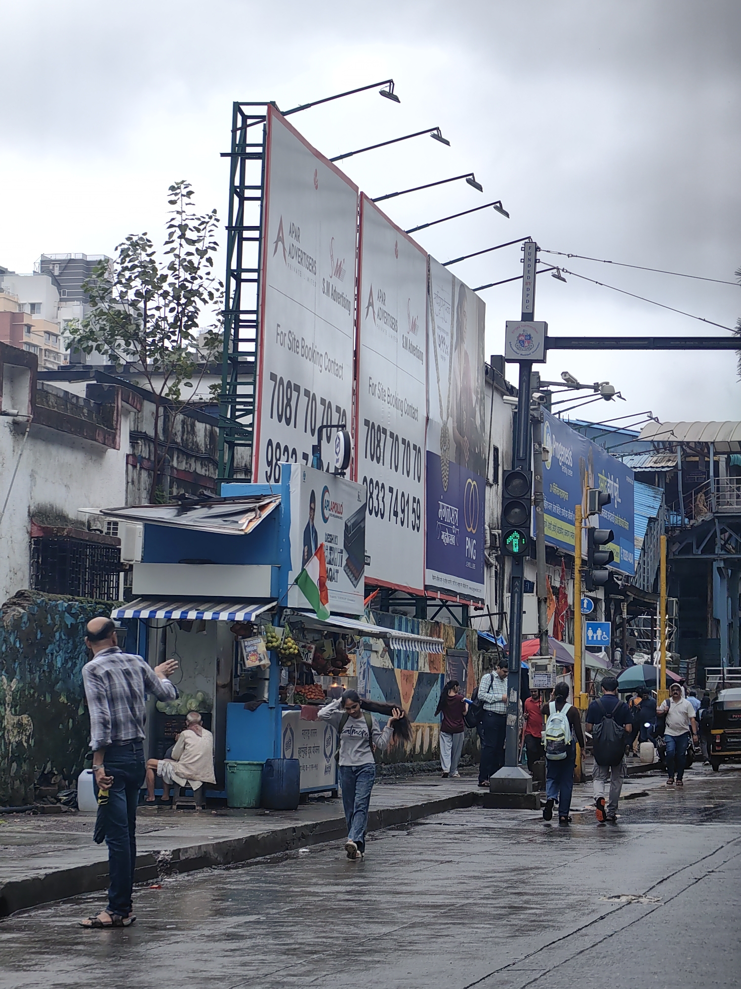 The image depicts a street scene in Mumbai, India, under an overcast sky. In the foreground, a man in a checkered shirt and jeans stands on the left side of the wet road, gesturing with his right hand. To his right, a vendor sits behind a stall selling fruits and juices, with a small Indian flag displayed. Several people are walking on the wet sidewalk and crossing the street, some carrying bags and umbrellas. A traffic signal shows a green pedestrian crossing light. Large billboards advertising "APAR ADVERTISERS" and "SM Advertising" are prominently displayed on the left side of the image, with phone numbers visible. Behind the billboards, apartment buildings are visible. The street is wet, indicating recent rain.