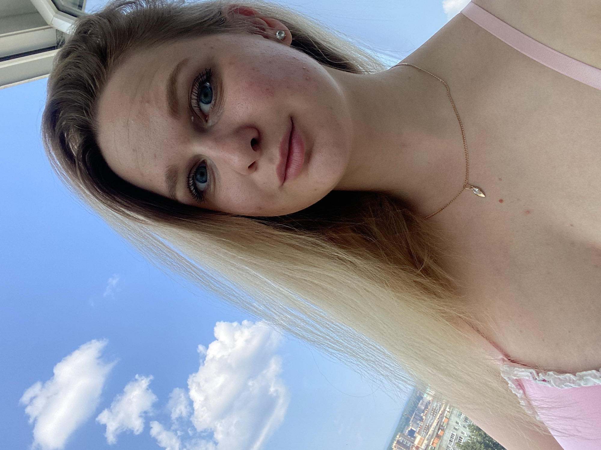 A young woman with blonde hair and blue eyes takes a selfie outdoors. She is wearing a pink top with lace trim and a delicate gold necklace with a small pendant. The background shows a bright blue sky with fluffy white clouds and a cityscape with buildings and trees in the distance. The woman's face is close to the camera, with her head tilted slightly. The image appears to be taken during the daytime, likely in the summer. The location is the city of Izhevsk, Russia.