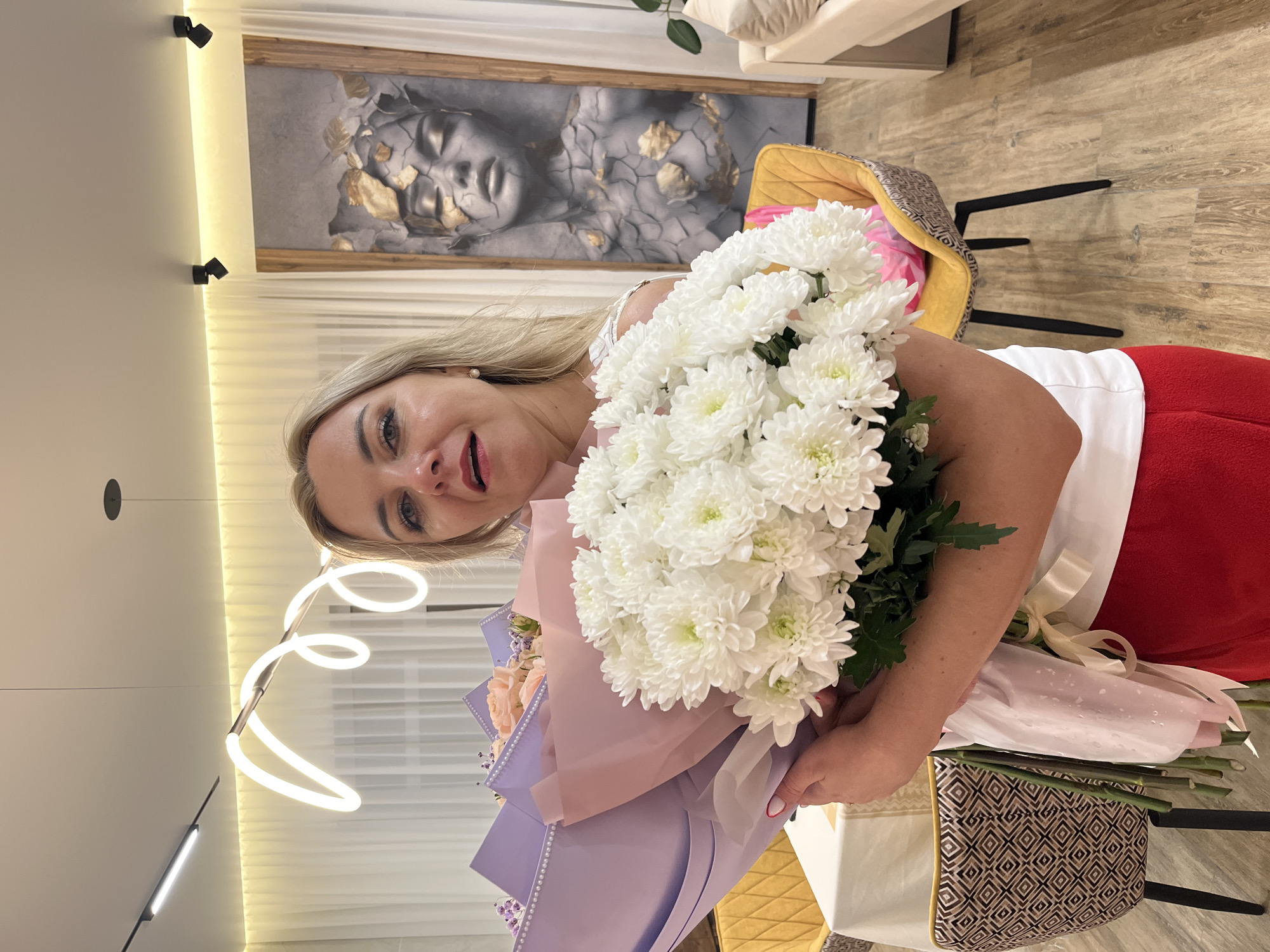 The image shows a blonde woman holding a large bouquet of white chrysanthemums and other flowers wrapped in pink and purple paper. She's smiling and looking at the camera. She is wearing a white top and red pants. In the background, there is a picture of a face with golden leaves. There is a sofa and chair visible. A neon light fixture hangs from the ceiling. The scene appears to be indoors, possibly in a home or apartment. The location is in Новая Чернушка, Russia.