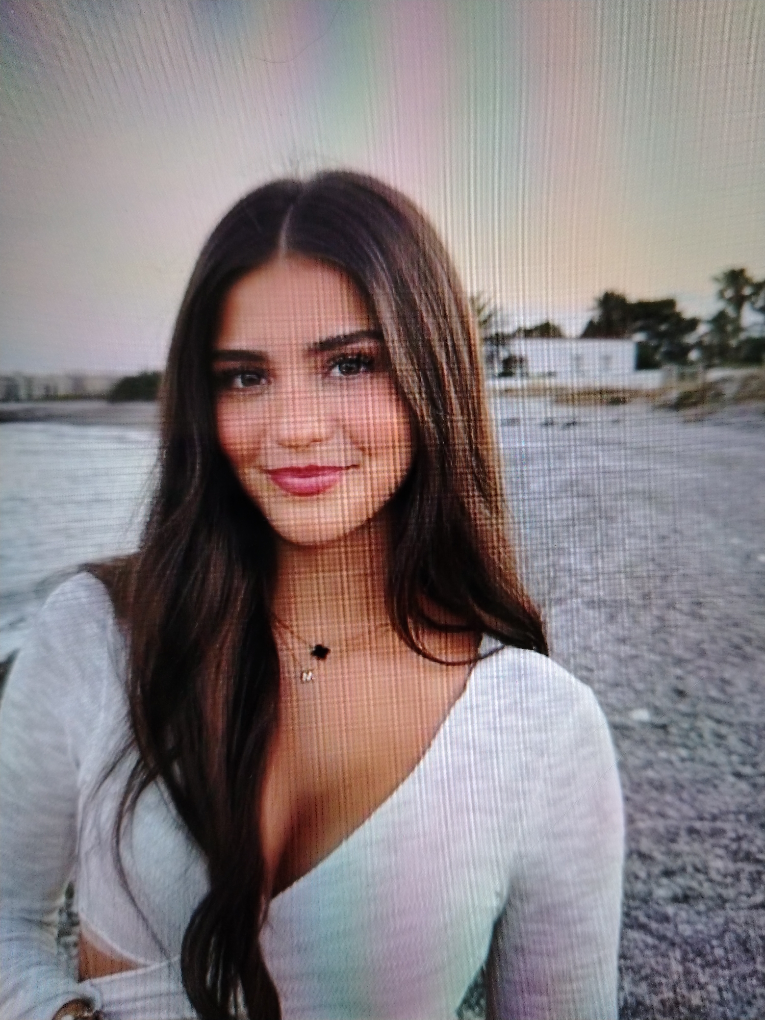 A medium shot captures a young woman with long, dark brown hair styled in a middle part. She is wearing a white, long-sleeved top with a deep V-neckline and a cropped hem, revealing a glimpse of her midriff. A delicate gold necklace with two charms, a black clover and a small initial "M," adorns her neck. Her makeup is subtle, emphasizing her dark eyes and rosy lips.
She stands outdoors, seemingly on a beach or a rocky shore. The background features a body of water, possibly the sea, stretching towards a distant horizon under a pale, diffused sky. A low-lying structure, possibly a building, is visible in the mid-ground, flanked by palm trees. The ground is covered with small, grey stones or pebbles. The lighting suggests it is either early morning or late evening.
This image is not located in Stavropol, Russia.