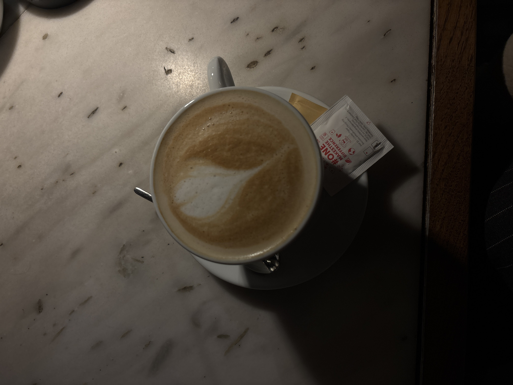 An overhead shot captures a cup of latte with a heart-shaped latte art design, resting on a white saucer on a light-colored, mottled marble tabletop. A metallic spoon is tucked under the cup's handle. Next to the cup, two sugar packets are visible; one is white and features the text "#ONE MAKES THE DIFFERENCE" along with recycling information in Italian and English, including "PAP 22 - Carta / Paper", "Raccolta differenziata / Recycling", "Non vendibile singolarmente. / Not to be sold separately", and the manufacturer "Fast Zuccheri Srl., Samarate (VA), Italia / Italy." The other packet appears gold or brown.
The scene is dimly lit, with a concentrated light source highlighting the coffee cup and immediate table area, casting prominent shadows to the right and bottom of the frame. The marble table surface shows dark speckles and veins, indicative of natural stone. A dark, wooden edge runs along the right side of the image, suggesting a table or counter, next to which dark, indistinct fabric (possibly clothing) is visible. The image captures a quiet moment of enjoying coffee, likely in a cafe or an indoor setting in Yerevan, Armenia. The overall mood is calm and contemplative due to the soft lighting and focus on the beverage.