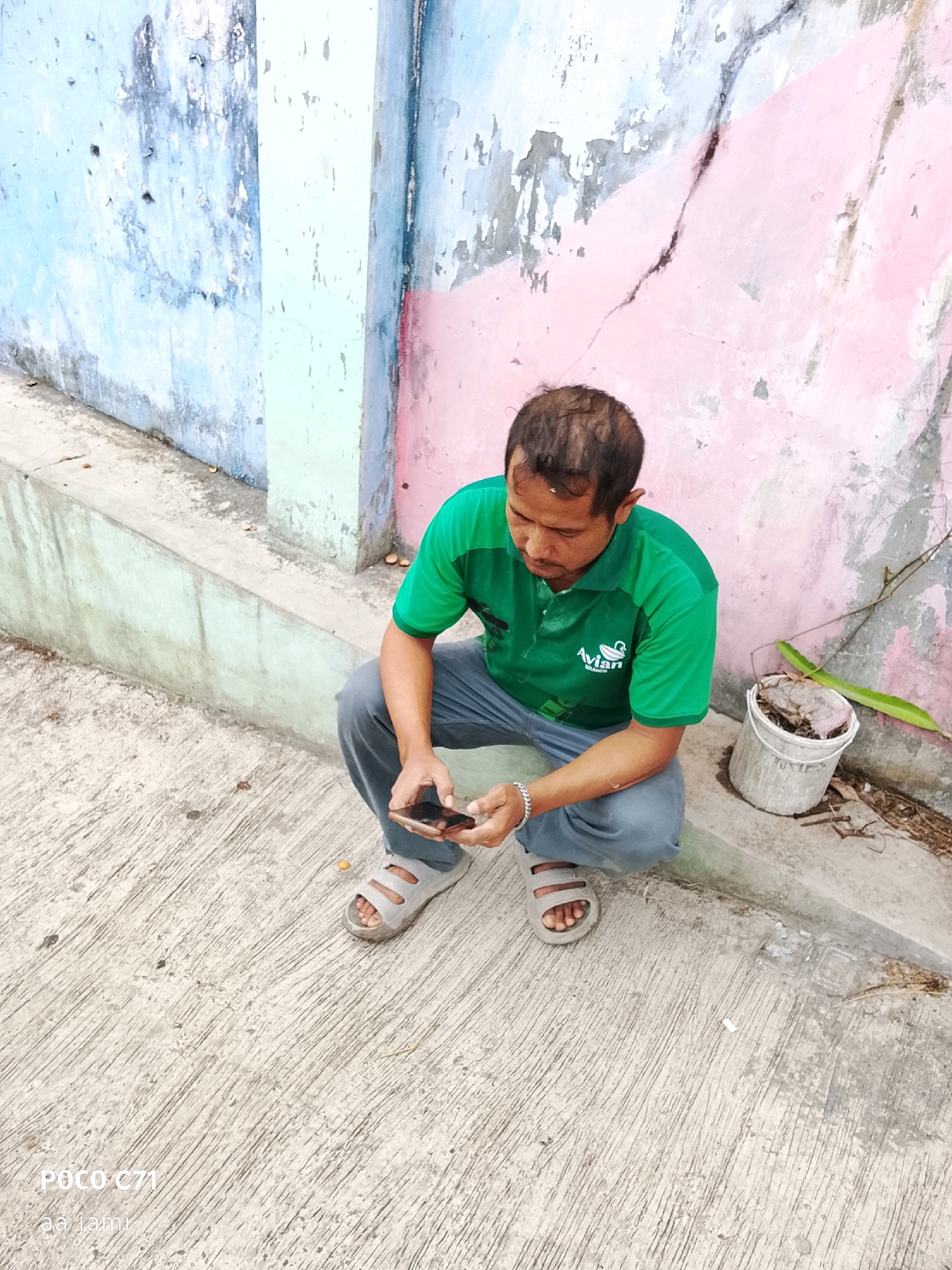 A man is crouching on a concrete ground, looking at his phone. He is wearing a green polo shirt with a bird logo and blue pants. He has on grey sandals. To his right, there is a white bucket with some debris in it and a long green leaf. The background is a cracked, painted wall with sections of blue, pink, and light green. The scene appears to be an outdoor area, possibly a street or alleyway, in Tasikmalaya, Indonesia, given the "POCO C71" watermark in the lower-left corner which is a brand of smartphone often used in Indonesia, and "aa jami" which may be a localized watermark.