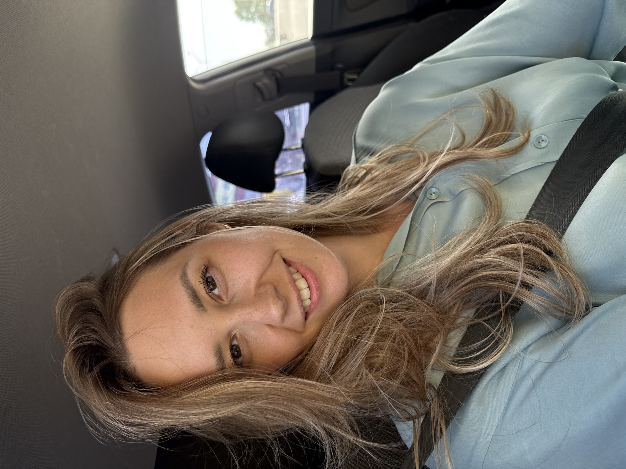 A young woman is inside a car, wearing a light blue button-down shirt and a seatbelt. She has long, wavy blonde hair and is smiling. The car's interior is visible, including the seat, headrest, window, and various car fixtures. The location is purportedly Chaikovsky, Russia, but there is no visual confirmation of this information in the image.