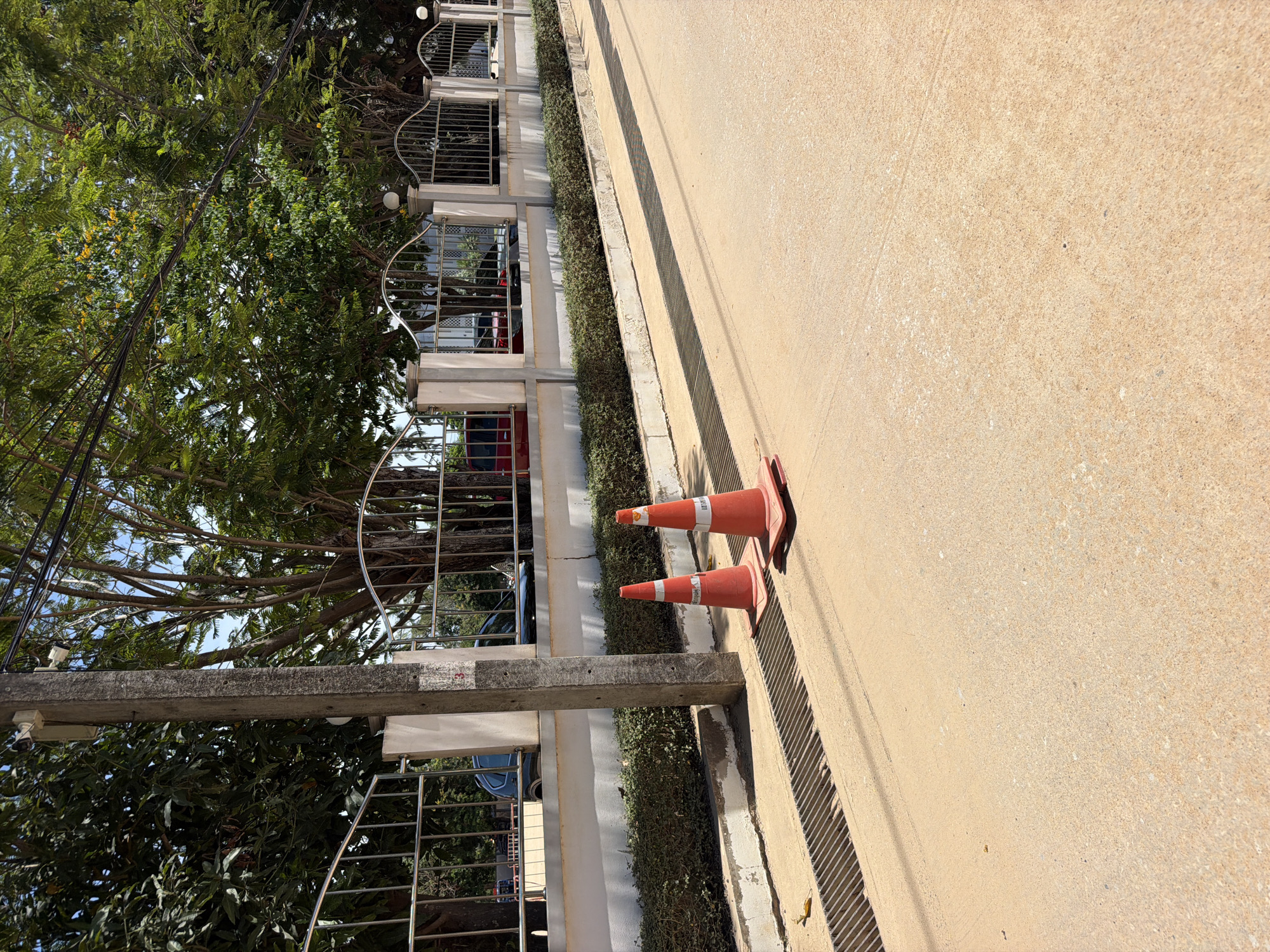 A bright, sunny day shot captures a paved road in Baan Bo Phut, Thailand, with a concrete sidewalk and a decorative metal fence in the background. Two orange traffic cones are positioned on the road, one slightly in front of the other. The fence runs along the back of the sidewalk, with lush green trees visible beyond it. A concrete utility pole stands to the left of the frame, near the fence. Cars are partially visible parked behind the fence. A metal grate, likely a drain cover, runs along the edge of the sidewalk and road. The overall scene is calm and devoid of human activity.