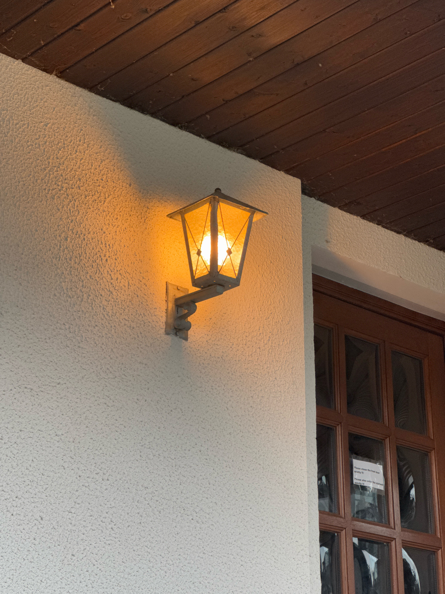 An exterior wall-mounted lantern with an illuminated bulb casts a warm glow on the textured, off-white wall of a building. The lantern is made of metal and glass and hangs from a decorative bracket. Above the wall, a wooden soffit with visible planks runs horizontally across the top of the image. To the right of the lantern, a wooden-framed door with multiple glass panes is visible. A small white sign with black text is affixed to one of the glass panes, which reads: "Please close the front door quietly !!!" and "Please also enter the stairwell quietly !!!". The scene suggests an entrance to a building, possibly residential or commercial, in the city of Cleves, Germany.