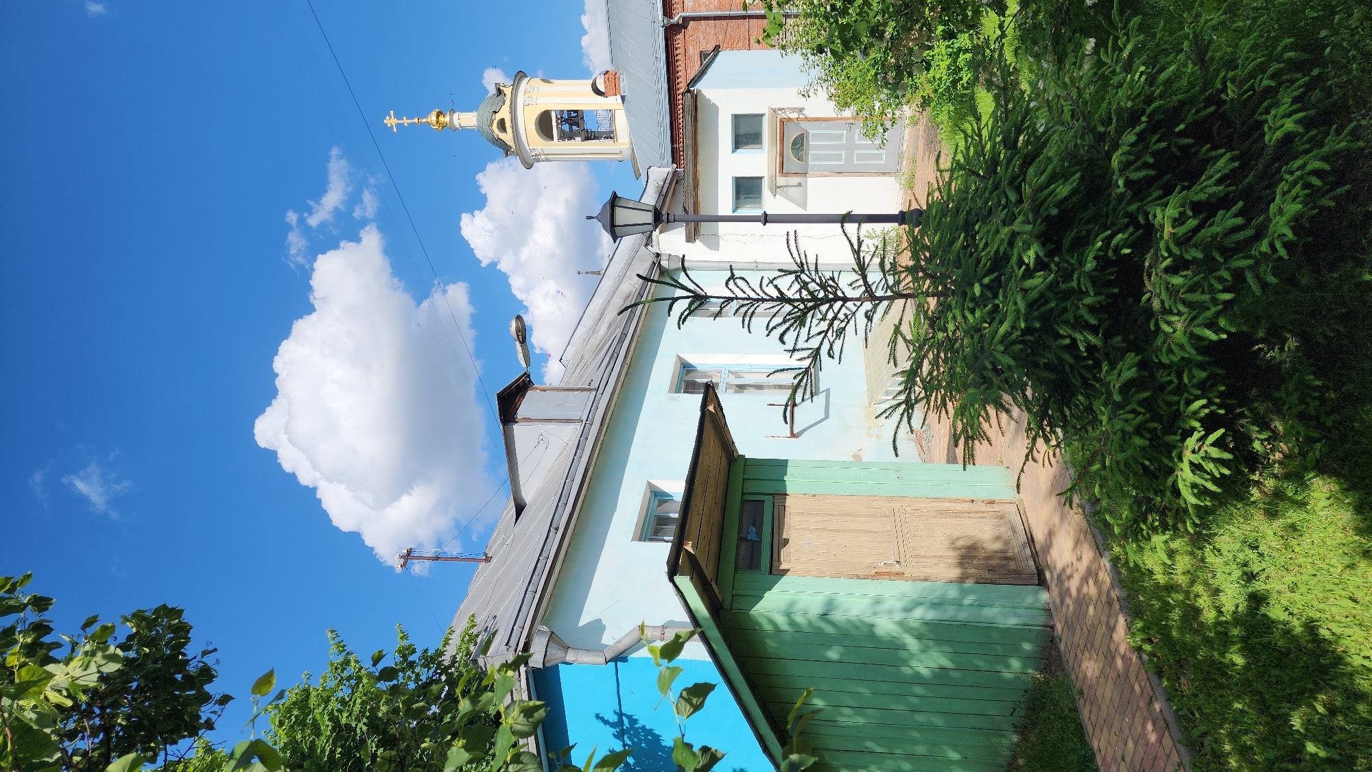 The image captures a vibrant, sunlit outdoor scene, looking up through a narrow passage between buildings towards a clear blue sky.
In the foreground and to the left, a traditional light blue house with a grey metallic roof is prominent. Its lower facade features a rustic dark green wooden fence or siding, with a wooden door recessed beneath a small canopy. Several windows with light blue frames are visible on the house's wall.
Adjacent to this, a narrow brick pathway extends from the foreground into the midground, bordered by a patch of bright green grass and a dense, dark green evergreen tree. A tall, black street lamp stands centrally, partially obscured by the foliage of the tree.
Behind the light blue house, another building with white walls, a light grey door, and visible windows can be seen, topped by a section of red brick. Above these structures, against a brilliant azure sky, the ornate golden dome and cross of a church bell tower rise, gleaming brightly under the sun.
The sky is a striking deep blue, adorned with several fluffy white cumulus clouds, indicating fair weather. The strong sunlight casts distinct shadows, suggesting it is mid-day. The architectural style, particularly the church steeple and the older wooden house, provides a glimpse into a historical urban landscape, consistent with the provided location context of Moscow, Russia. The scene conveys a peaceful and bright atmosphere. No people or visible text are present.