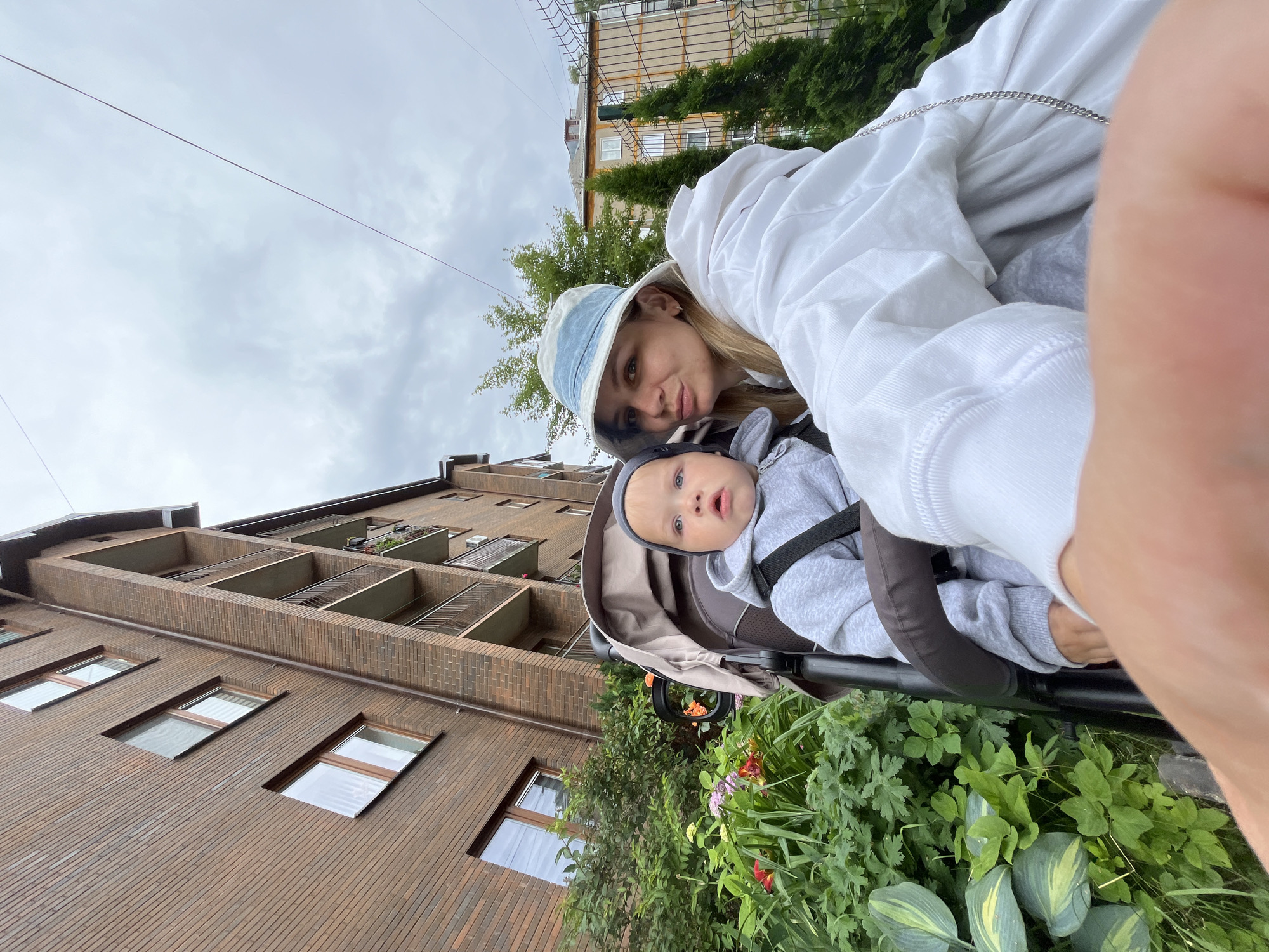 In Pionersky, Russia, a woman wearing a white hoodie and a blue and white bucket hat is captured in a selfie with a baby. The baby is seated in a stroller and is wearing a grey hooded sweatshirt and a grey beanie. They are positioned in front of a multi-story brick building with balconies. Lush green foliage and flowers are visible in the foreground, adding a touch of nature to the urban setting. The sky above is overcast, suggesting a cloudy day. A metal fence can be seen in the background, indicating a boundary or enclosure.