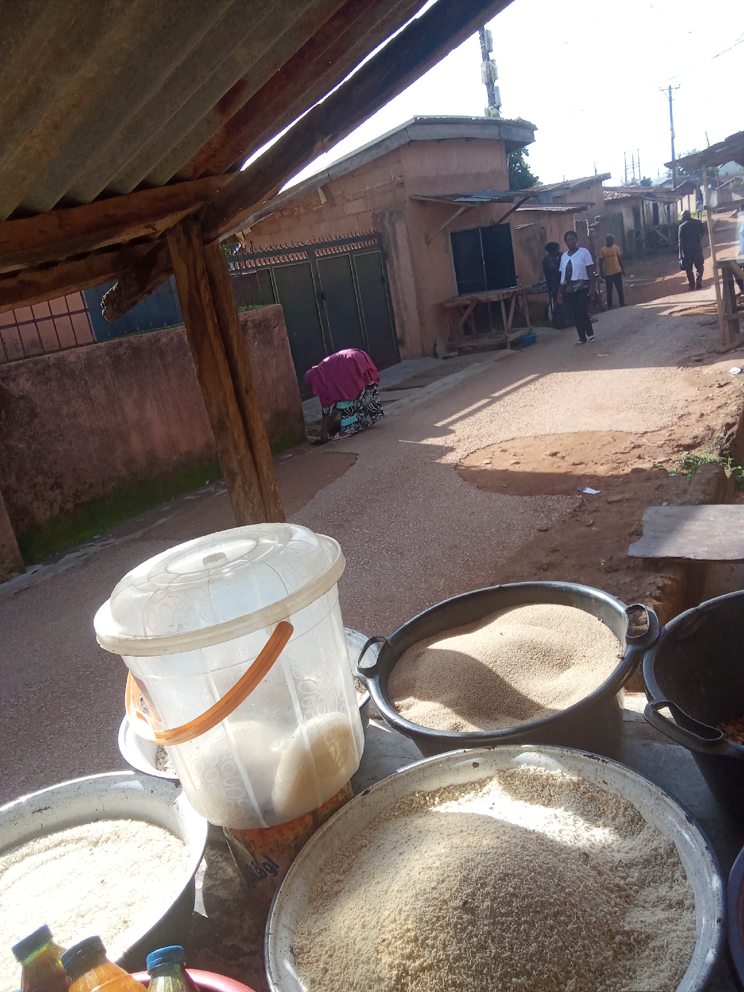 This is an outdoor scene in the city of Jos, Nigeria, likely a market or roadside stall. The foreground features several large metal basins filled with what appears to be granular goods, possibly rice or grain, and a large clear plastic bucket with a handle, also containing a granular substance. In the background, there are buildings with corrugated iron roofs and brick or concrete walls. Several people are visible in the midground, walking along a dusty path. One woman in a purple and patterned outfit is bent over, appearing to be working or attending to something on the ground. Another woman in a white t-shirt and dark pants is standing and looking towards the viewer. A man in a dark shirt and pants walks away from the camera. The scene is illuminated by natural sunlight, casting shadows on the ground and buildings. The overall impression is one of a bustling daily life scene in a Nigerian urban setting.
