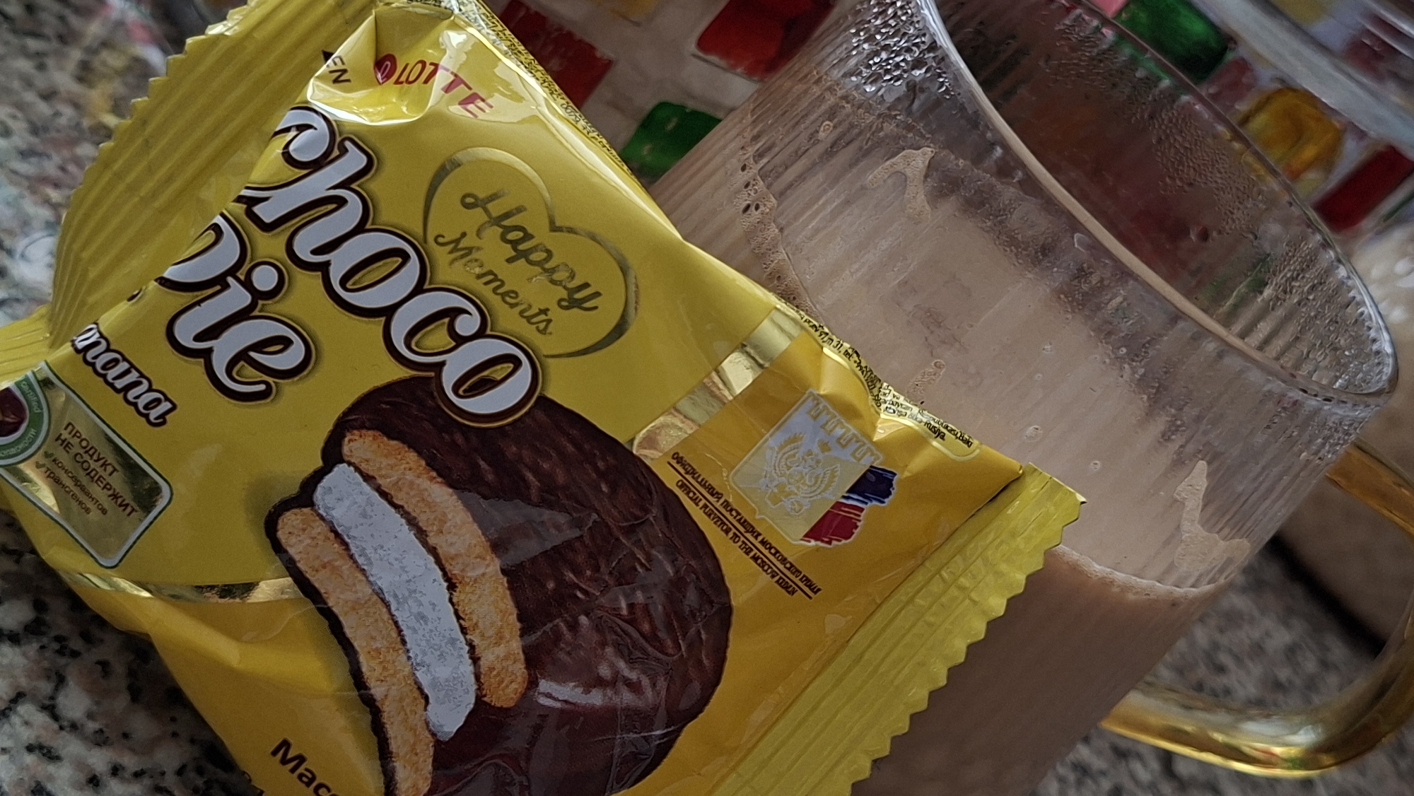 A close-up shot shows a yellow wrapper with the words "Happy Moments Choco Pie" and an image of a chocolate-covered pie. The wrapper is partially open, revealing a bite taken from the pie, showing its layers. A glass filled with a light brown liquid, possibly coffee or milk, is visible in the background, with condensation on its exterior. The glass has a golden handle. The overall setting appears to be indoors, with a textured surface underneath. The image is not specific to Myadzyel, Belarus.