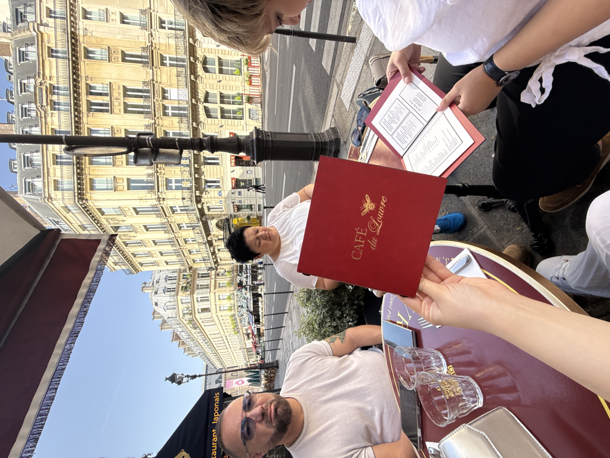 The image shows a group of people sitting at an outdoor cafe in Paris, France. Two people are visible at the table, a man wearing sunglasses and a woman. Both are wearing casual clothing (T-shirts) and are sitting at a maroon-colored table. The table has glasses and a metal container and one person is holding a red menu from "Café du Louvre". Another person is holding a white menu. In the background is the street, crosswalk, other diners, and a building facade typical of Parisian architecture. A black restaurant awning is on the lower left of the picture.