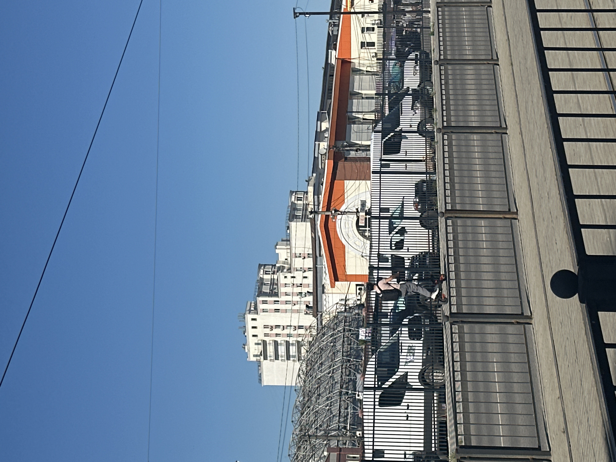 This is an urban street scene captured on a clear, sunny day. The image is composed of a bright blue sky at the top, a series of buildings in the midground, and a paved walkway and road in the foreground, separated by a fence.
In the upper portion of the frame, a vast, unblemished blue sky is visible, with several power lines traversing horizontally across it.
The midground features a dense array of city buildings. To the left, several multi-story residential or office buildings, primarily white or light-colored, stand tall, some with visible balconies and numerous windows. Towards the center, there's a row of commercial-style buildings, characterized by their reddish-orange roofs or architectural details. Below these buildings, a large, complex metal structure, possibly scaffolding or a decorative installation, is prominent.
The foreground is dominated by a wide, paved pedestrian area or walkway on the right, which casts long, distinct shadows from a sturdy metal fence that runs along its length. These shadows suggest strong overhead sunlight. To the left of this walkway, beyond the fence, is a paved road with visible white lane markings. Several cars are parked or driving on this road, partially obscured by the fence. On the paved walkway, a single person is visible, riding a scooter or a small bicycle. A large circular shadow, likely from an overhead street lamp or similar object, is cast on the far right of the walkway.
No discernible text is visible on any signs or buildings to indicate a specific location. The time of day appears to be midday due to the high sun and sharp shadows. The overall impression is one of a bustling city environment under bright, clear weather conditions.