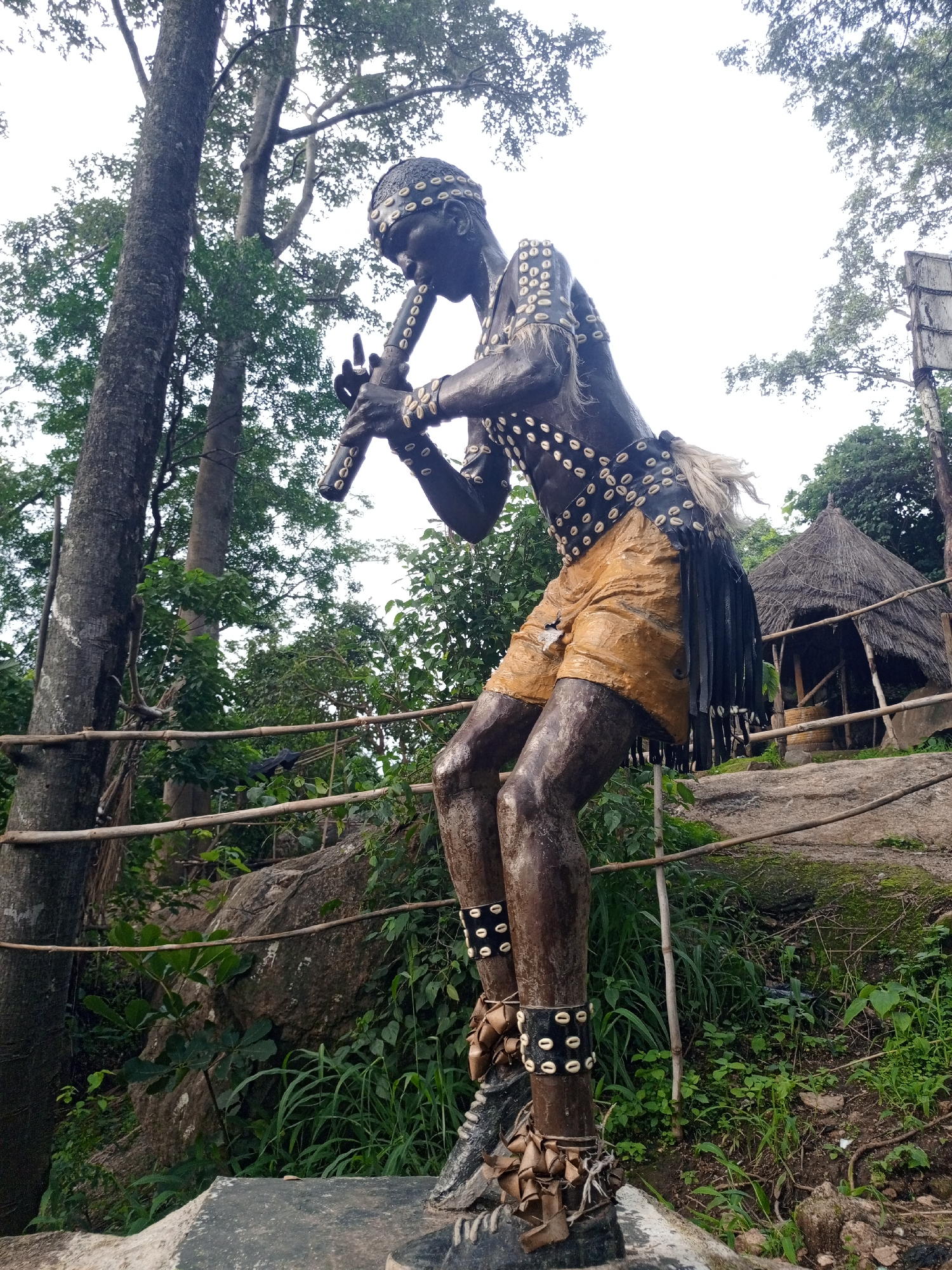 This image depicts a bronze sculpture of a musician playing a flute, positioned outdoors in what appears to be a natural or park-like setting, likely in Jos, Nigeria. The sculpture is detailed, showing intricate decorative elements on the figure's clothing and body, including what seem to be shells adorning a headband, chest, and armbands. The figure is wearing a headdress and a loincloth or shorts, with a fringed belt. The background features lush green vegetation, trees, and a thatched-roof structure that resembles a hut or shelter. A bamboo railing is visible in the foreground, suggesting a pathway or viewing area. The overall environment suggests a cultural or historical exhibition.