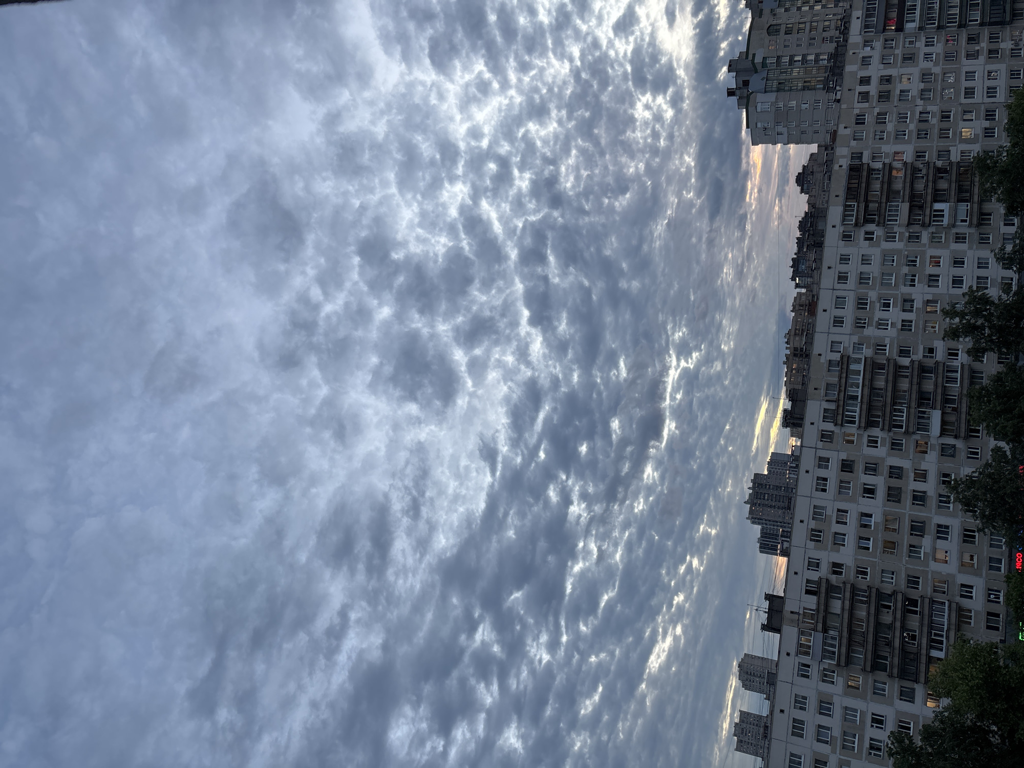 A dramatic, overcast sky fills the top two-thirds of the frame, with a pattern of grey and white clouds stretching across the horizon. Below the sky, a long row of apartment buildings stretches across the width of the image. The buildings are multi-story structures with numerous windows and balconies. In the foreground, the tops of dark green trees are visible. The scene appears to be an urban landscape at dusk or dawn, with the muted lighting and cloud cover creating a somber mood.