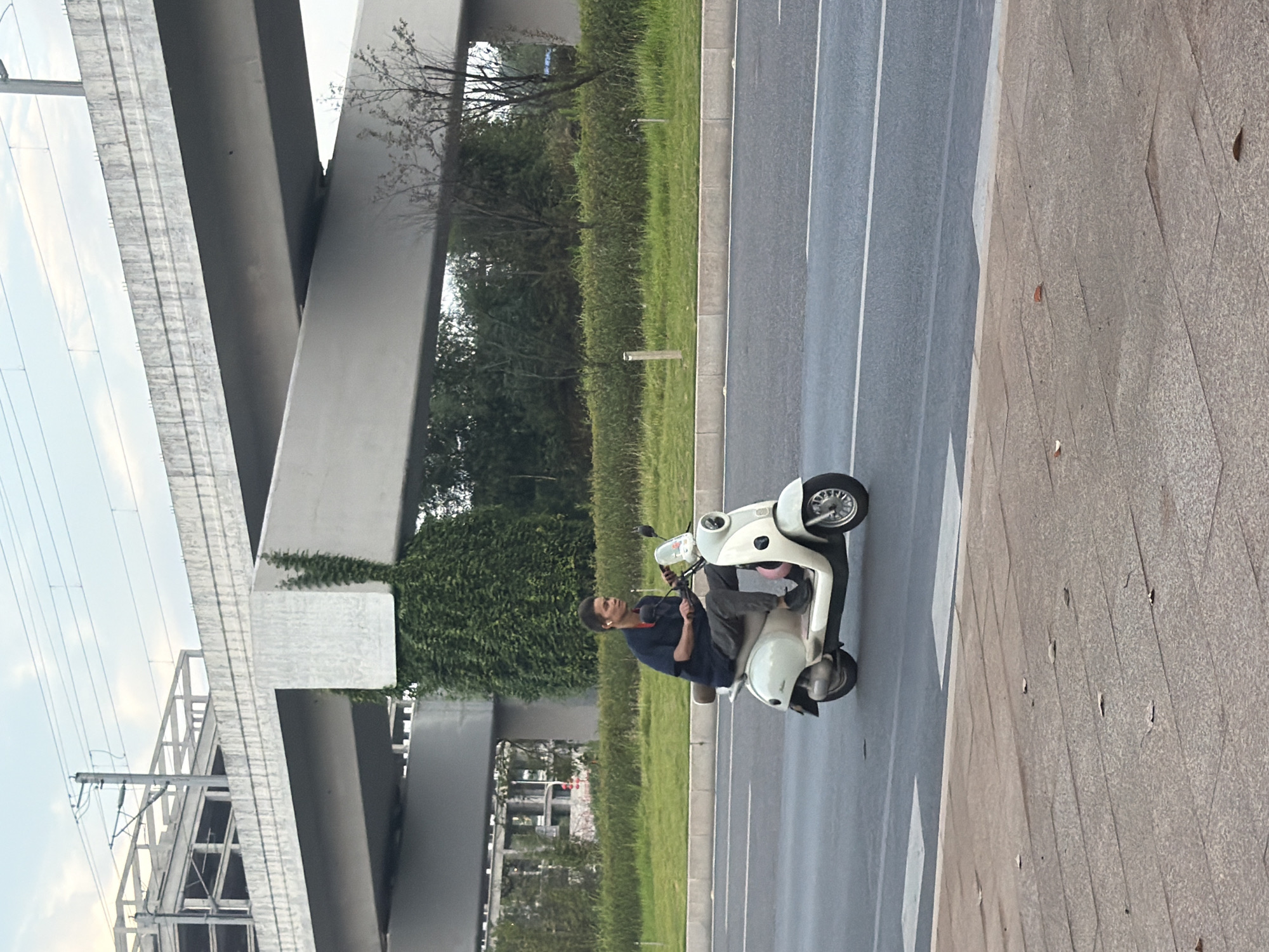 A man rides a cream-colored scooter on a paved road in Jiangjin, China. He is wearing a dark blue short-sleeved shirt and gray pants, with white earbuds in his ears. The scooter has a pink object on the rear storage area. The road has white lane markings and a pedestrian crosswalk in the foreground. Behind the road is a green grassy area with bushes and trees, separated by a concrete barrier. In the background, a large elevated concrete structure, possibly an overpass or part of a building under construction, is visible with electrical wires running overhead. The sky is overcast.