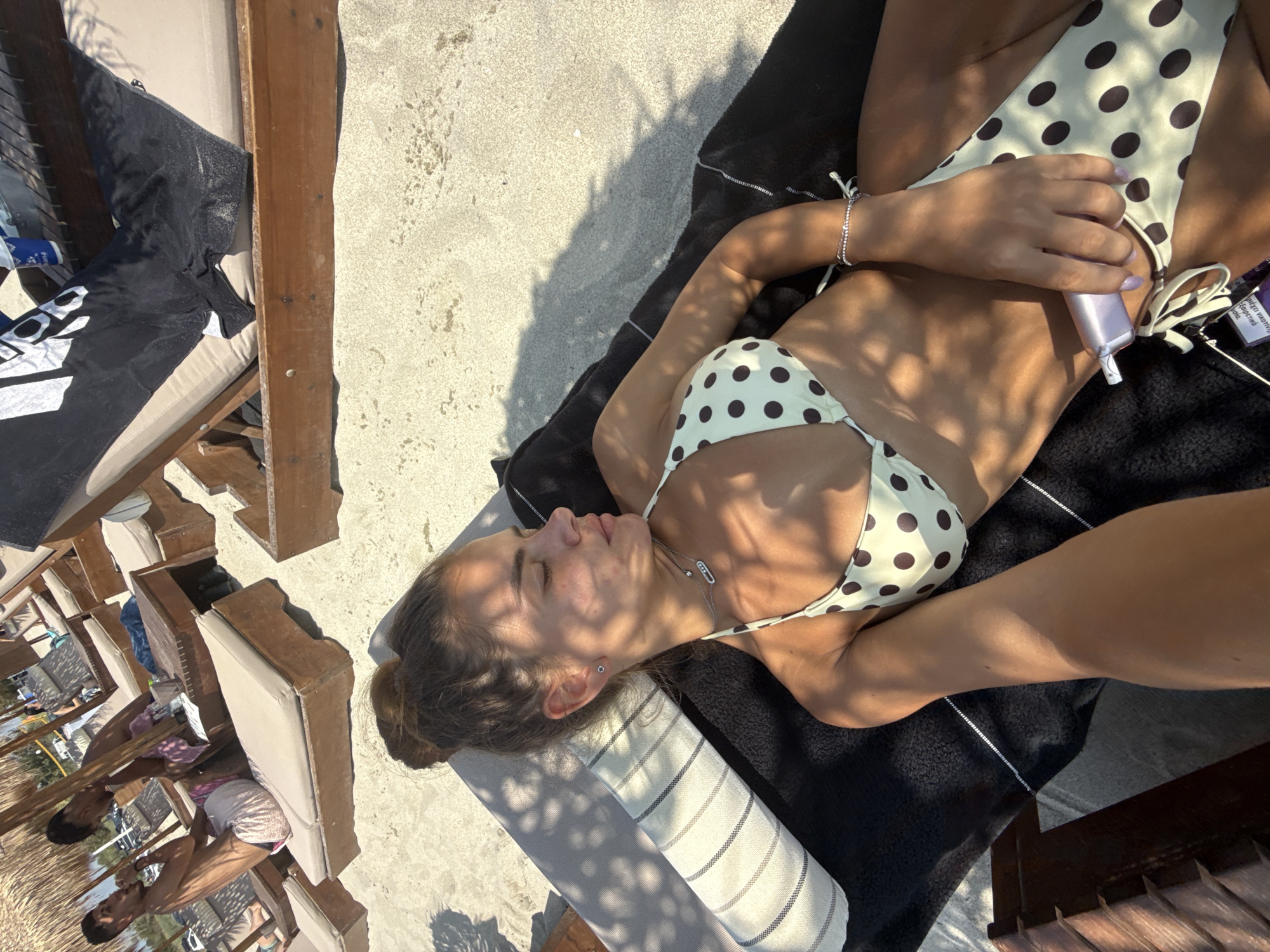 A young woman with her eyes closed is lying on a lounge chair on a sandy beach in Katerini Municipality, Greece. She is wearing a light-colored bikini with dark polka dots and a silver necklace. Her hair is pulled back in a bun. She is holding a small, light-purple object in her right hand. The background shows other lounge chairs, beach umbrellas, and people in the distance. The lighting suggests it is daytime, and shadows from the umbrellas create patterns on the sand and the woman.
