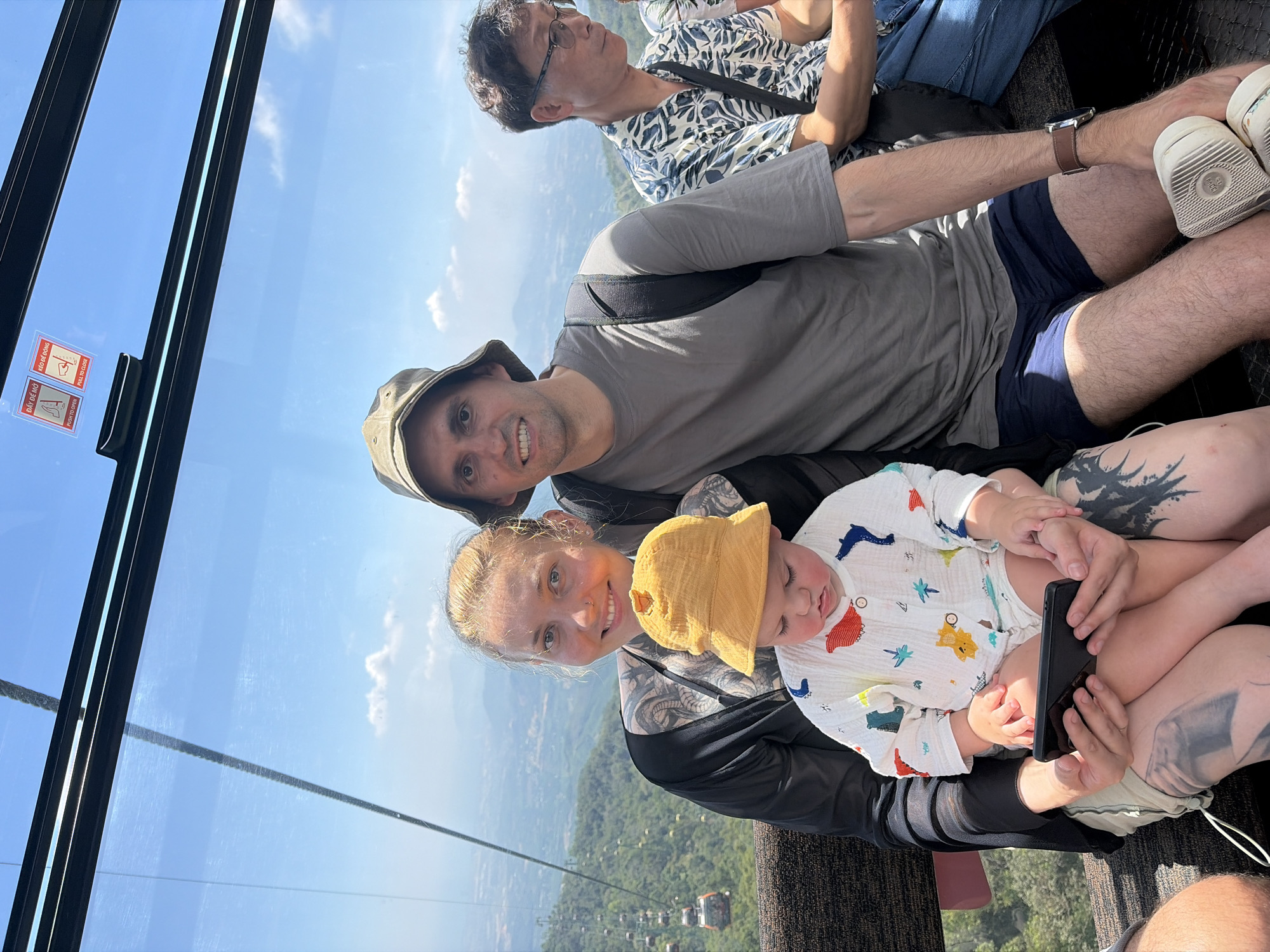 A medium shot shows four people in a cable car in Đà Nẵng, Vietnam. A family of three is seated on the lower half of the frame, while an elderly person is sitting in the upper half. The foreground shows a man wearing a gray shirt and a khaki-colored bucket hat, a woman in a black jacket, and a baby wearing a yellow bucket hat and patterned white onesie. The woman holds a black smartphone in her hands. The foreground also shows parts of two other people, one wearing shorts, tennis shoes, and a watch and the other wearing jeans and a button-down shirt. Cable car cables and green landscape with many small cable cars are visible in the background.