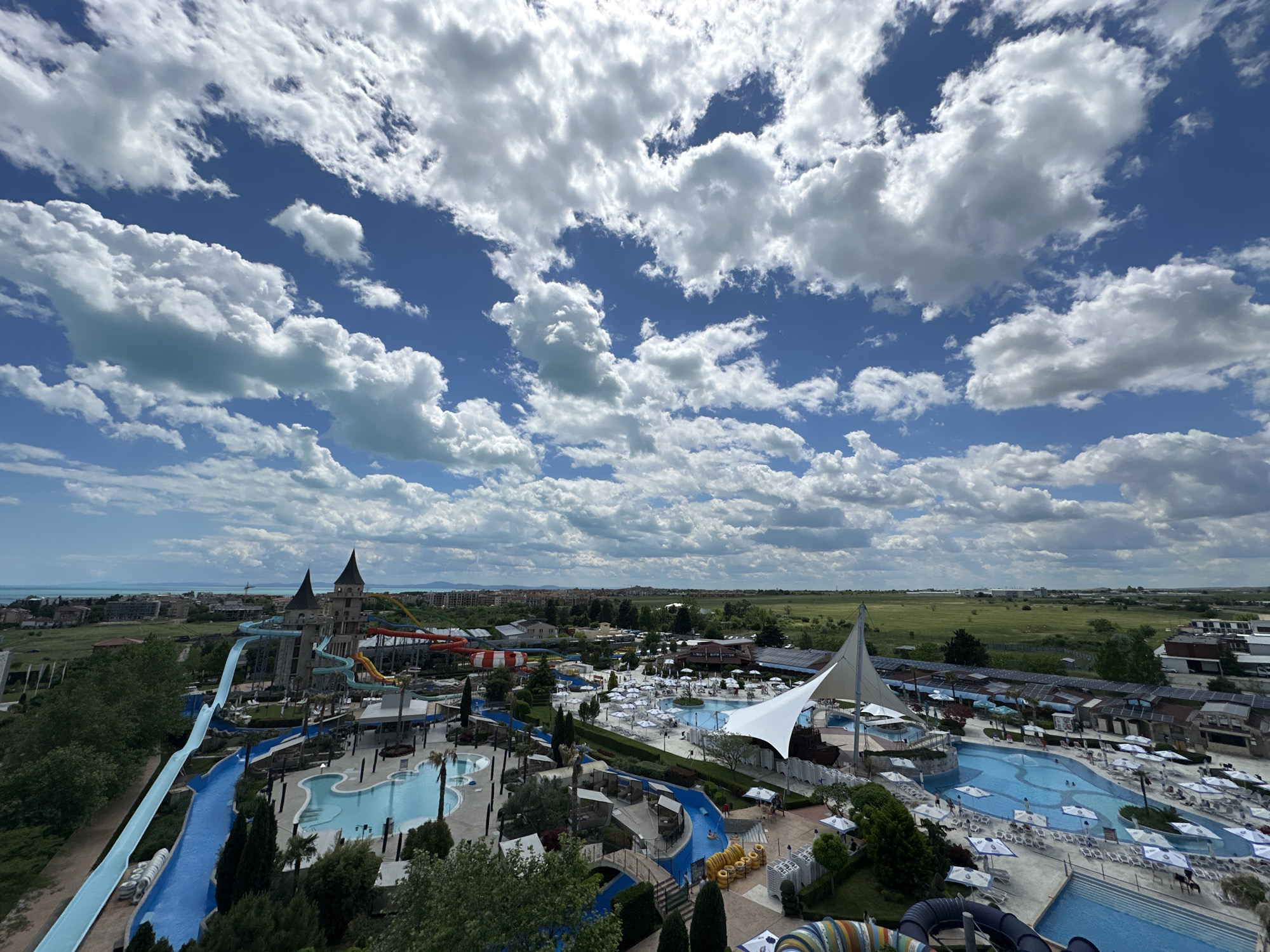 The image shows a water park in Ravda, Bulgaria, under a sky filled with white, puffy clouds and blue sky. In the foreground, there are several pools of various sizes and shapes, connected by blue water slides. Some of the pools have people swimming in them, while others are surrounded by white sun umbrellas and lounge chairs. Landscaping features including trees and bushes are also visible. In the midground, there are a variety of buildings, including a structure with pointed turrets and other entertainment facilities. In the background, there is a view of the Black Sea, with some buildings and vegetation near the coast. Beyond that is a grassy field.