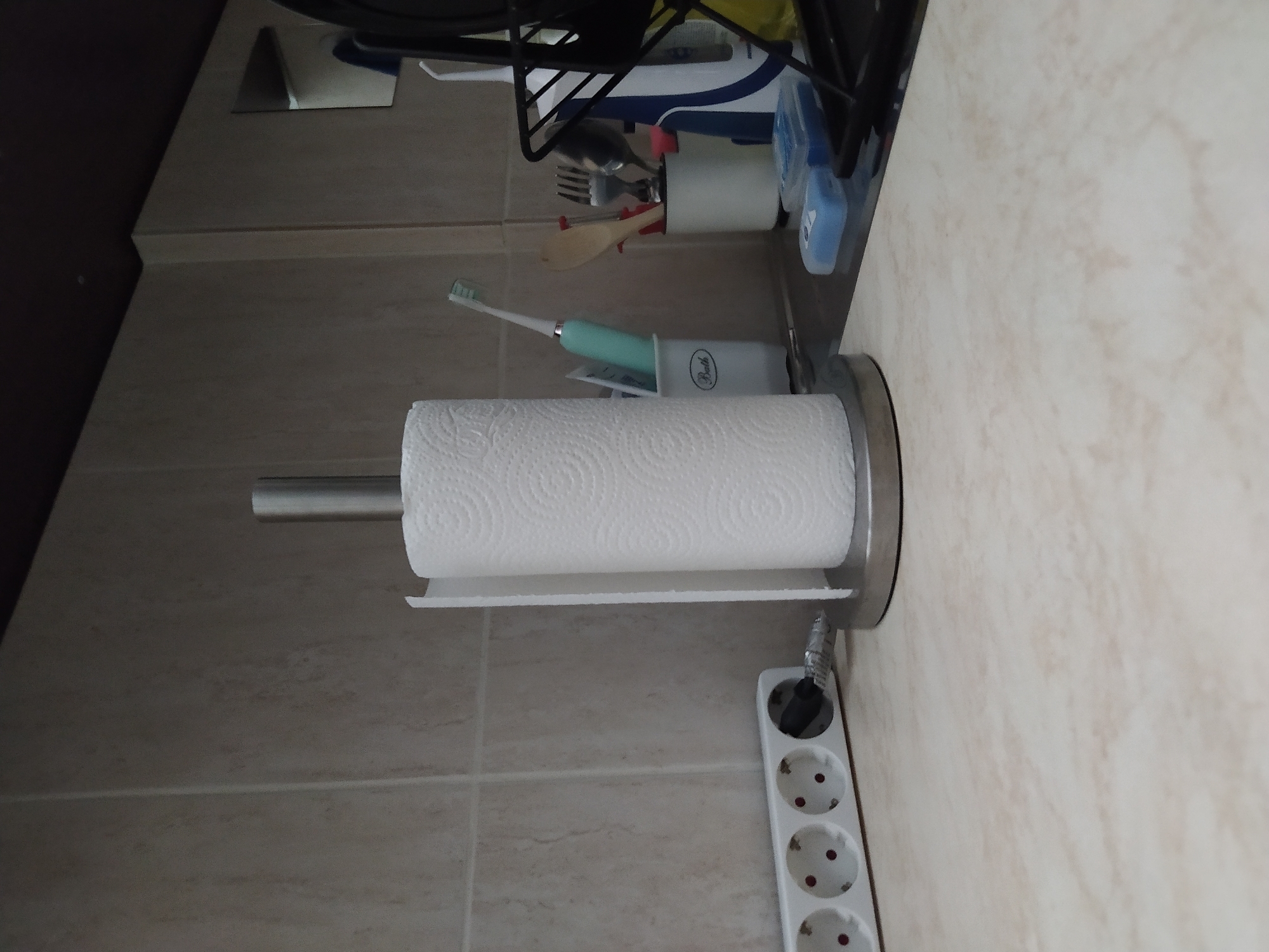 The image shows a kitchen scene. A roll of white paper towels sits on a silver-colored holder attached to a light-colored tile wall. An extension cord is visible beneath the paper towel holder. Further up the wall, a toothbrush and holder, along with kitchen utensils in a container, are present. The location is not determinable from the image, and therefore I cannot comment on the city of Sofia, Bulgaria.