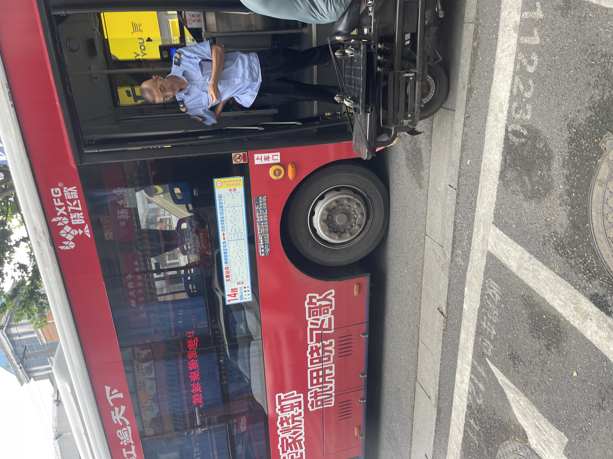 A red and white bus with Chinese characters, including "XFG" and "晓飞歌" (Xiaofeige), is stopped at an open door on an urban street in the city of Unknown, Unknown. A male bus driver with a light buzz cut, wearing a light blue short-sleeved uniform shirt with dark blue epaulets and black trousers, stands at the open front door, leaning against the door frame. He is looking down at a small white object held in his hands. His left hand rests on his hip. Visible through the bus window are interior elements and a yellow sign with a partially visible shopping cart icon and "YOU." A sign displaying the route number "14" and a route map is affixed inside the bus near the door. The bus displays "家烧虾" (home-style braised shrimp) and "就用晓飞歌" (use Xiaofeige) on its lower red side panel. To the right of the bus, the rear section of a black three-wheeled scooter or utility motorcycle with a flat cargo platform is parked on the sidewalk. The asphalt road in the foreground features white painted lines and the reversed alphanumeric sequence "0E22L1" written on the pavement. A concrete sidewalk is visible between the road and the bus/scooter. In the background, a building with a blue-tiled roof and green foliage is partially visible under an overcast sky, with overhead cables traversing the scene.