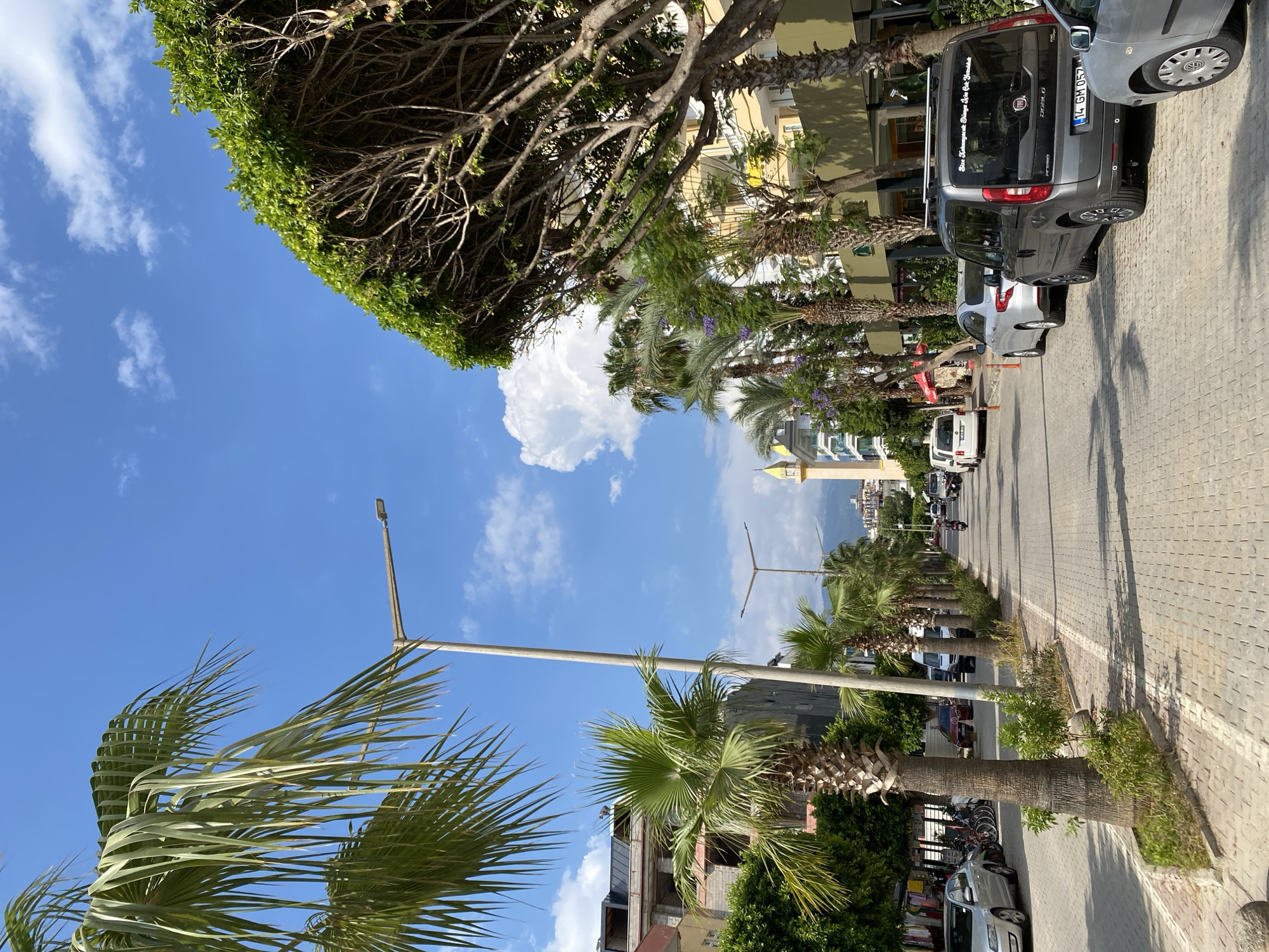 The image depicts a street scene in Alanya, Turkey, under a clear blue sky with scattered white clouds. The perspective is from a low angle, looking up towards the sky and across a street lined with palm trees and buildings.
In the foreground, the fronds of a palm tree sway gently, suggesting a breezy day. A street lamp is visible, indicating an urban or suburban environment. The street itself is paved with cobblestones and lined with parked vehicles, including a grey Fiat Doblo with a license plate reading "14 GM 057" and other cars and motorcycles. Buildings with yellow and green facades are situated along the street, some appearing to be under construction. The presence of a mosque minaret in the distance provides a strong cultural cue of the location.
The time of day appears to be daytime, likely morning or afternoon, given the bright sunlight casting shadows. The overall atmosphere is that of a typical day in a warm climate, with the lush greenery of the palm trees contrasting with the urban elements. There are no discernible people in the scene. One of the cars has text on its rear window, but it is not legible enough to be transcribed.