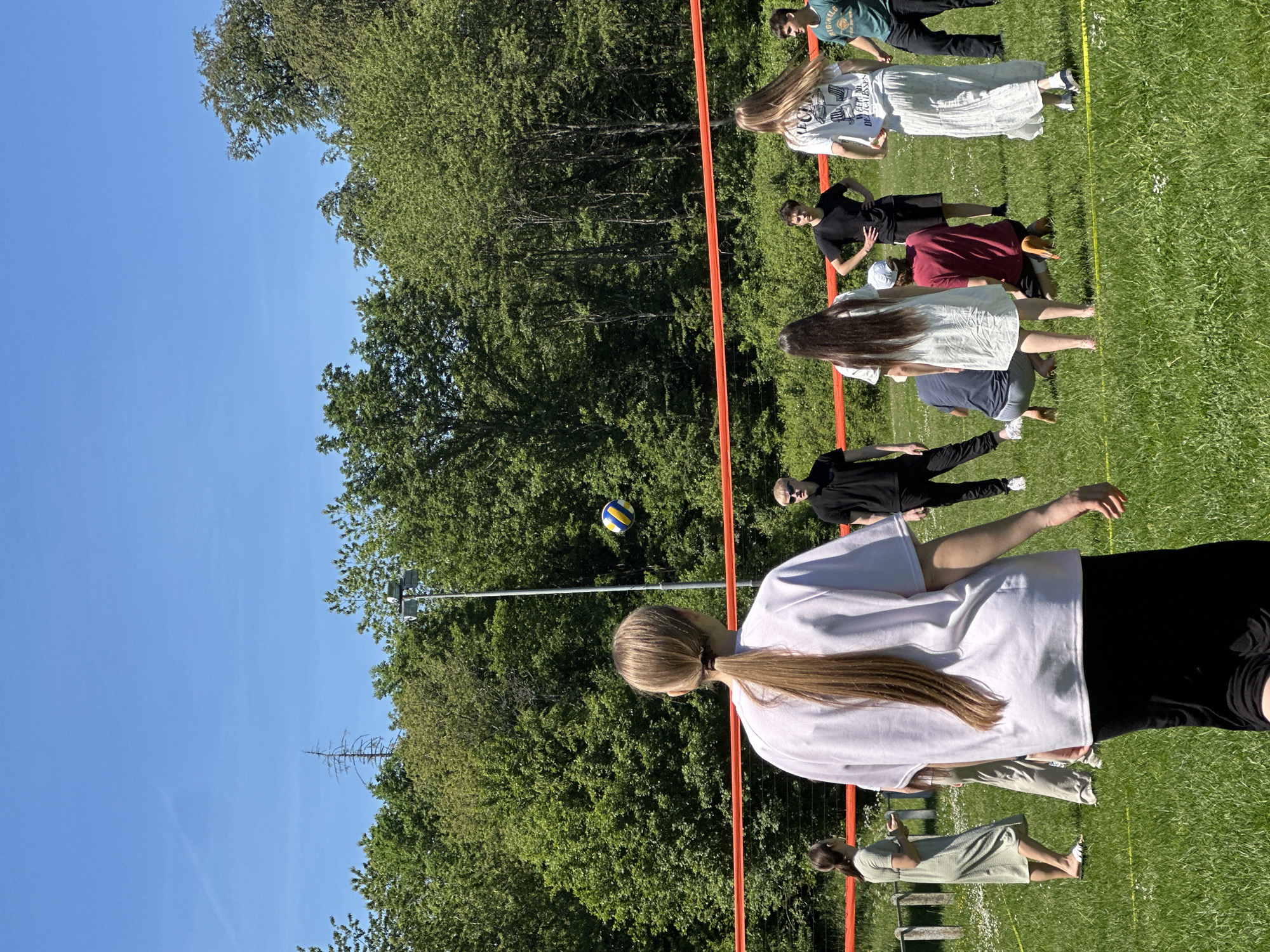 The photograph shows a volleyball game taking place on a grassy field in Grünberg, Germany. The sky is clear and blue. A group of at least eight people, a mix of genders and ages, are visible on the field. Some are positioned near the orange net, while others are further back. A volleyball is suspended in mid-air above the net. The surrounding environment includes a backdrop of dense green trees. A utility pole is visible in the background. The surface of the field is mostly green grass. The volleyball net is supported by a metal pole. Yellow lines are painted on the field.
