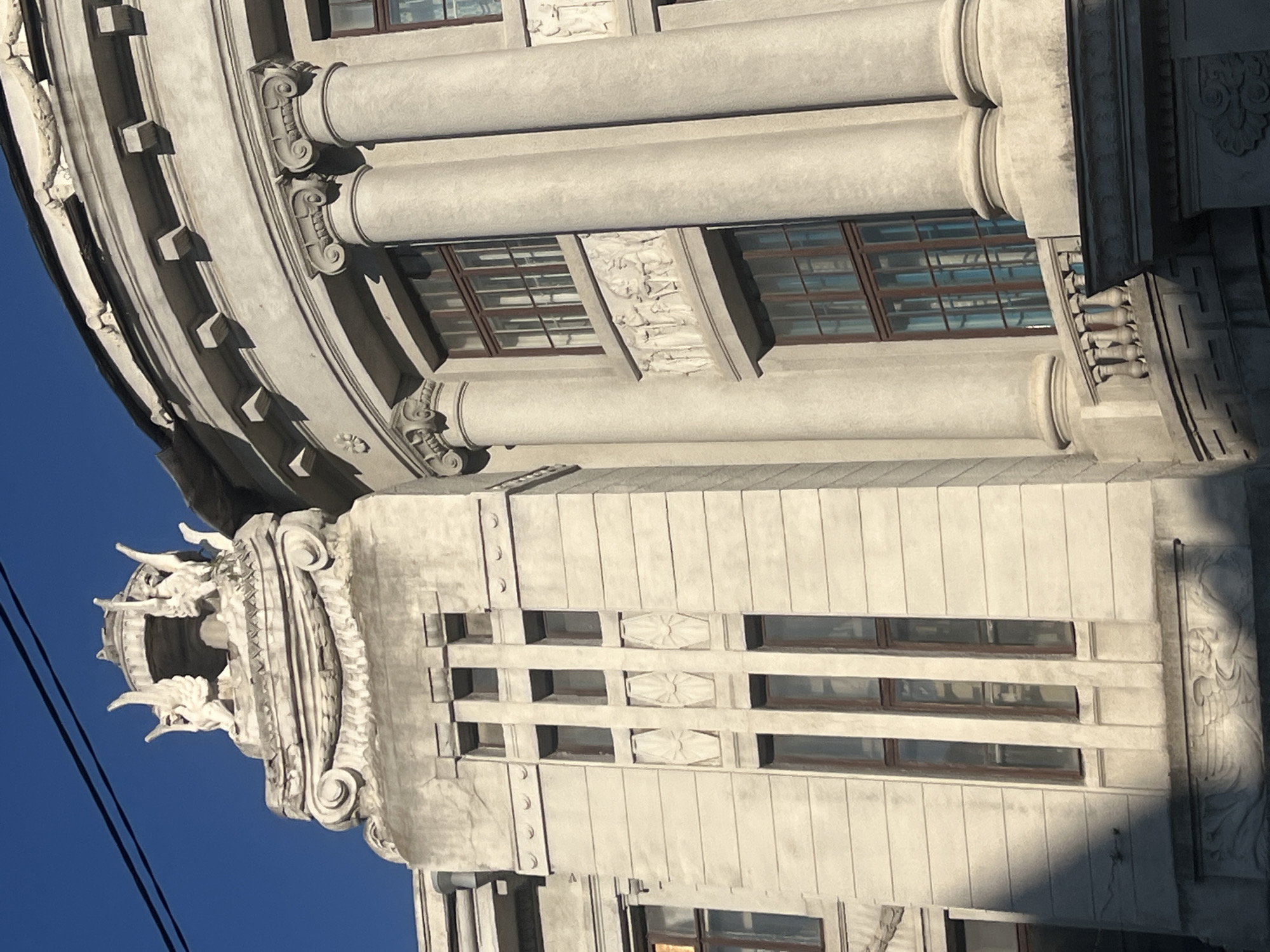 The image shows the facade of an Art Nouveau building, possibly the former Kharkiv Trade Union Building, located in Kharkiv, Ukraine. The building features intricate architectural details, including a curved section with classical columns and a more angular section with vertical windows. Decorative elements such as sculptural figures and bas-reliefs adorn the exterior. The sky is clear and blue, indicating fair weather. The building's architecture suggests a historical and culturally significant structure.