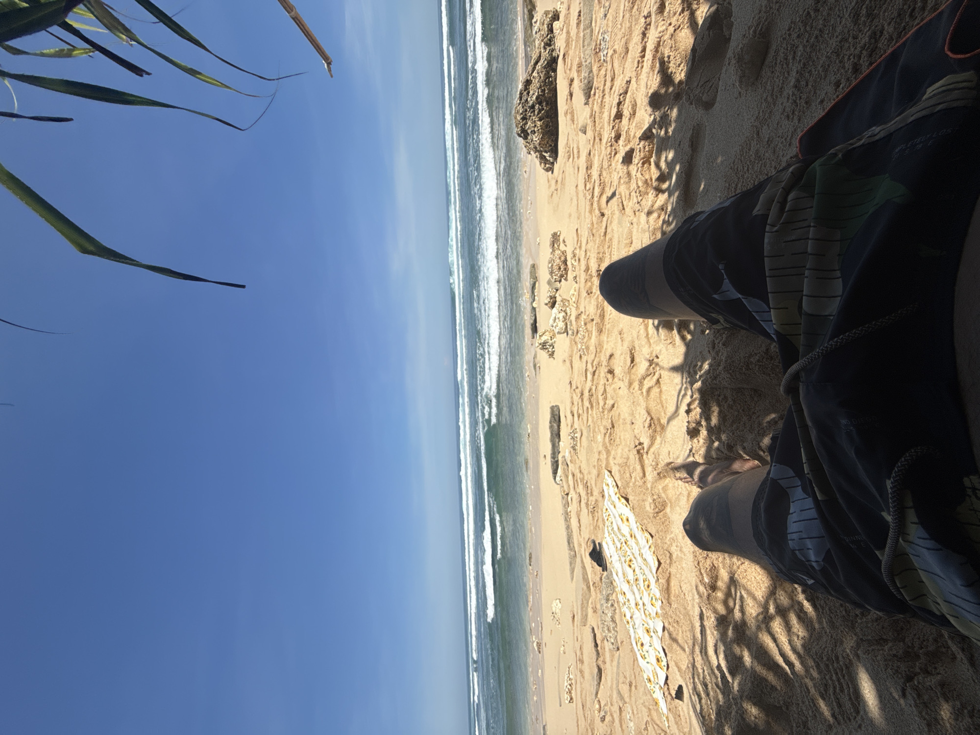 The image depicts a person sitting on a sandy beach in Kuta Selatan, Indonesia, with a view of the ocean. The person is wearing dark swim trunks with a light-colored print, and their legs are visible. A patterned towel lies near their feet on the sand. The ocean has visible waves breaking along the shoreline. In the background, the sky is clear and blue. Palm leaves are partially visible in the upper left corner of the image. The sand is golden-brown. A rock is visible between the beach and ocean. The scene appears to be taken during daylight hours.