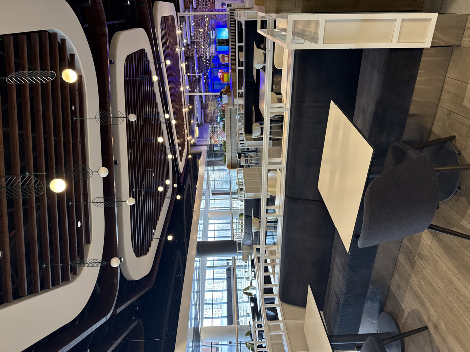The image depicts a spacious, modern dining area or food court located in the Blockbuster Mall in Kyiv, Ukraine. The scene is largely unoccupied, viewed from a low angle across multiple seating arrangements.
In the immediate foreground, two dark blue upholstered booth sections are visible, each paired with a square, light-colored table supported by a black central pedestal. A dark grey fabric chair with black legs is positioned at the right-hand table, with a similar chair partially visible on the left. The floor is covered in light wood-effect planks.
Beyond the foreground seating, a white slatted partition separates dining zones. Further into the midground, more dark blue booths, individual light-colored tables, and chairs are arranged. The area extends towards large, clear windows on the left, revealing exterior multi-story buildings under an overcast sky.
In the distant background, primarily to the right, is a food service or entertainment zone characterized by a textured brick wall and several brightly illuminated counters in blue and other colors. One individual, an adult, is faintly visible near these counters, facing away from the camera. No specific actions or interactions are discernible for this person.
The ceiling features a complex, multi-layered design with dark wooden beams, undulating white soffits, and numerous suspended light fixtures. Many of these fixtures consist of spherical white bulbs housed within decorative, dark metal frames resembling abstract leaves. Other light sources include simpler white globe pendants and recessed spotlights, uniformly illuminating the space. The overall environment is clean, contemporary, and well-lit.