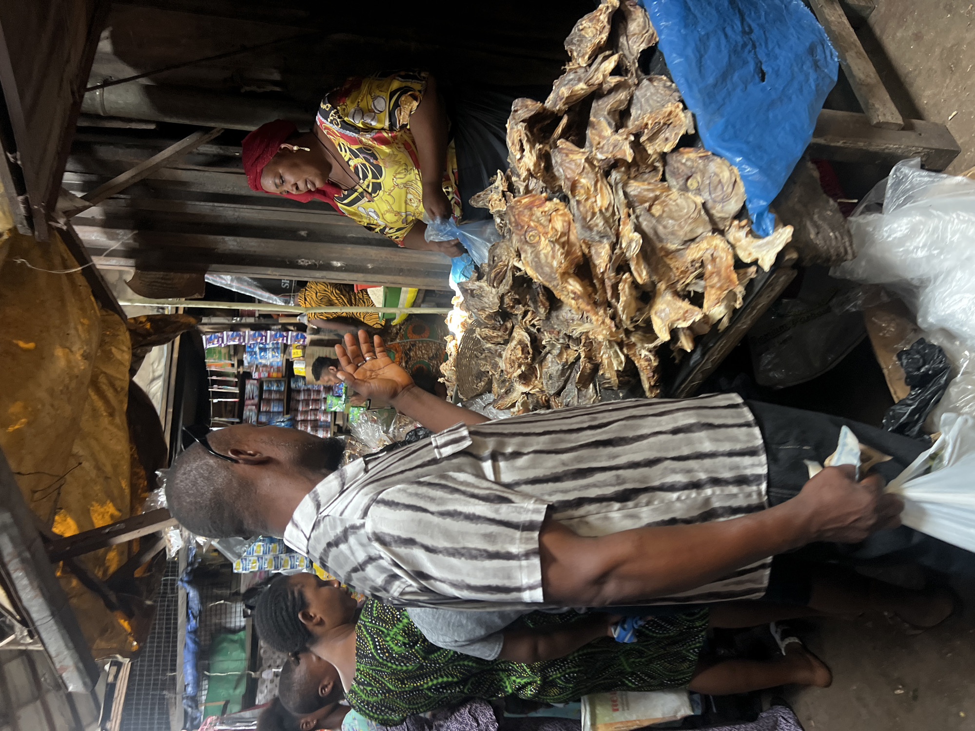 In Nkpor, Nigeria, a marketplace scene unfolds with an array of dried fish prominently displayed. A man, wearing a striped shirt, faces a woman at a stall, gesturing with his hand while holding a white plastic bag. The woman, adorned in a vibrant yellow and red patterned top and a red headscarf, engages with him. Behind them, shelves are stocked with various goods, and the general ambiance suggests a bustling commercial hub. The ground is littered with debris, common in such environments.