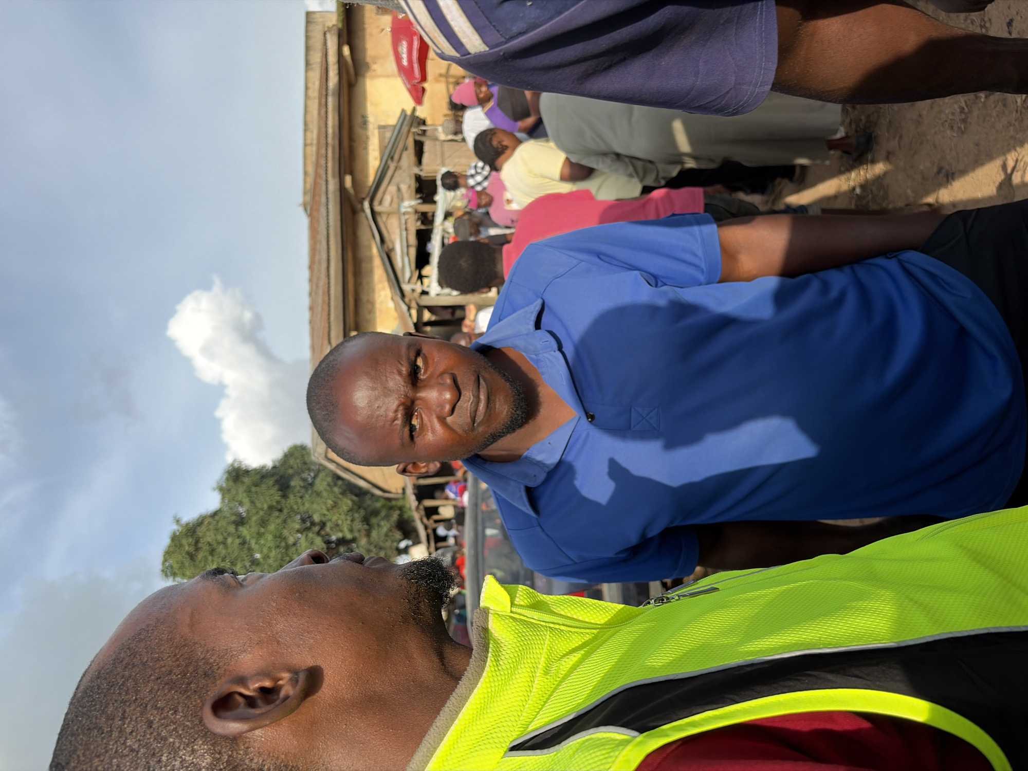 In Gura Top, Nigeria, a medium shot captures a group of people in an outdoor setting under a cloudy sky. In the foreground, two men are prominently featured. The man on the left, facing away from the camera and slightly to the right, wears a bright yellow vest over a dark garment, possibly with red accents. His skin tone is dark, and he has a close-cropped hairstyle. To his right, the man is facing the camera directly, his expression serious, with a closely-trimmed beard and short, dark hair. He is wearing a blue polo shirt. Behind these two individuals, a crowd of people is visible, many wearing colorful clothing, suggesting a community gathering or market. The background includes structures that appear to be market stalls or small buildings with corrugated metal roofs, indicative of a typical Nigerian urban or semi-urban environment. A red umbrella is also visible in the middle ground, adding to the market atmosphere. The ground appears to be dirt. The lighting suggests it is daytime, with sunlight filtering through the clouds.