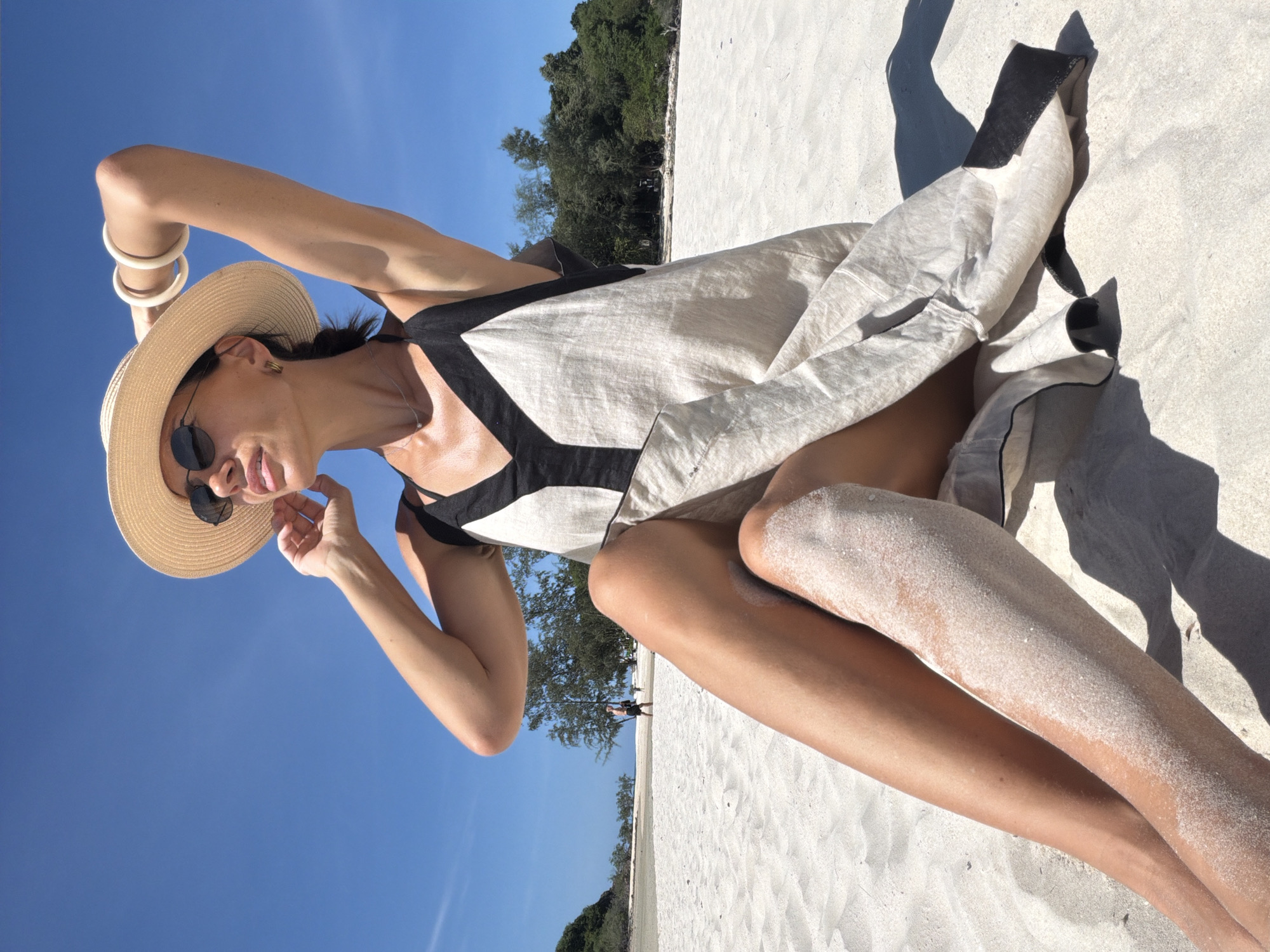 A woman sits on a white sandy beach in Baan Plai Laem, Thailand, with her legs crossed and one arm raised above her head, adjusting a straw hat. She wears sunglasses, a light-colored dress with dark trim, and white bracelets on her wrist. The sun shines brightly, casting shadows on the sand, and lush green trees line the background. The ocean is visible in the distance, with a person walking along the shoreline. The sky is a clear, vibrant blue.