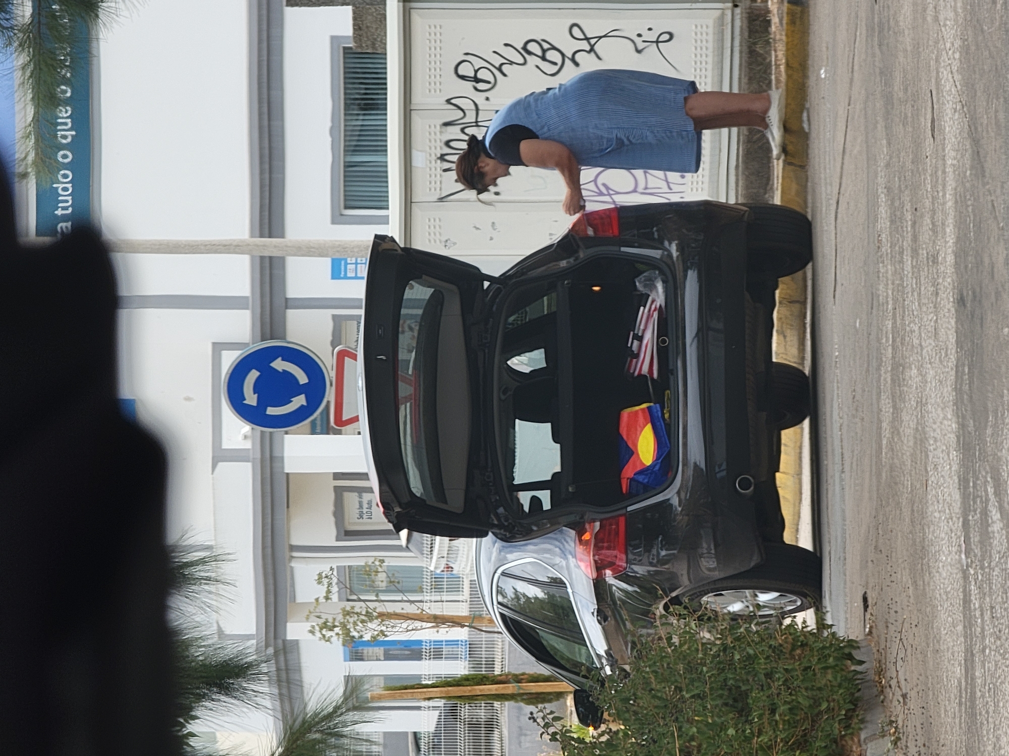 The image depicts an outdoor scene in Loulé, Portugal, with a black car parked on a street. The car's hatchback is open, revealing items inside, including a red, yellow, and blue flag or cloth, and what appears to be a bag with red and white stripes. A woman, wearing a blue and white striped dress and white shoes, is standing next to the rear of the car, facing away from the viewer and appearing to be engaged with the contents of the trunk. She has her hair pulled back. In the background, a white building with grey trim is visible, featuring windows and what looks like a sign with text. A blue circular traffic sign with white arrows indicating a roundabout is also present on a pole near the building. Graffiti is visible on the white surface behind the woman. The ground is asphalt, with a yellow curb running along the edge of the street. Green foliage is in the foreground to the left, partially obscuring the car. The sky is not visible.