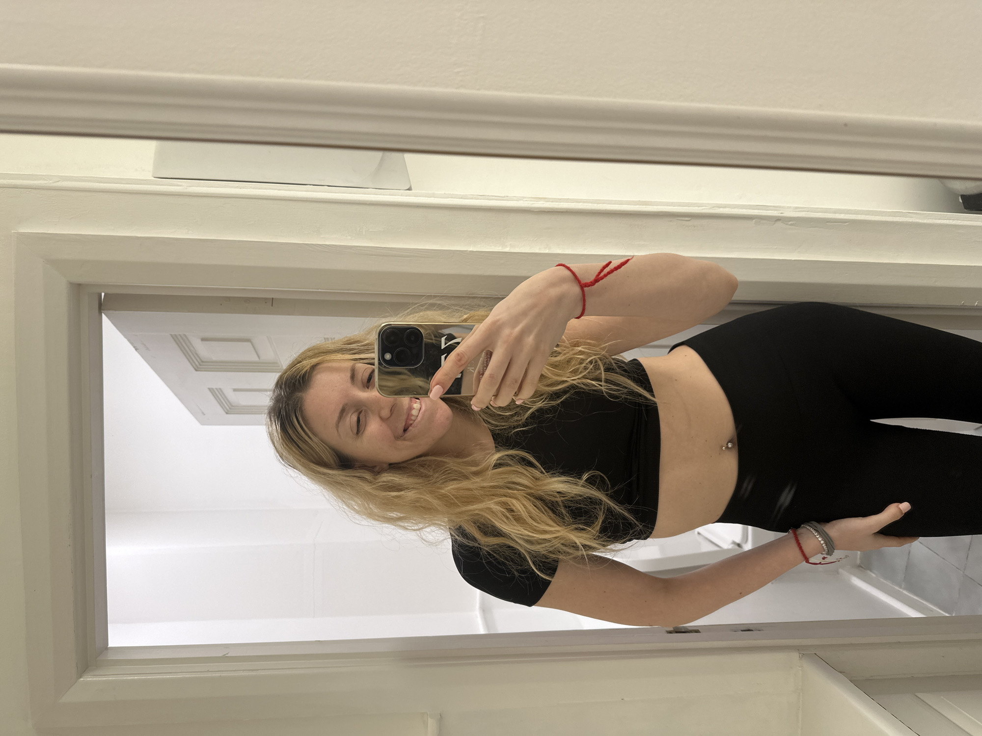 The image displays a female subject capturing a mirror selfie indoors. The subject has long, wavy blonde hair, is smiling with her left eye partially closed, and holds a dark-cased smartphone in her right hand, partially obscuring the lower right side of her face. She wears a black short-sleeved crop top and black leggings, revealing a silver barbell piercing in her navel. Her left wrist is adorned with multiple bracelets, including red string and beaded designs, while her right wrist has a single red string bracelet. The environment is characterized by white walls and white doorframes. A doorway behind the subject opens to an interior space featuring light-colored tile flooring and a partially open white paneled door. No specific visual identifiers are present to determine the precise location within London, United Kingdom.