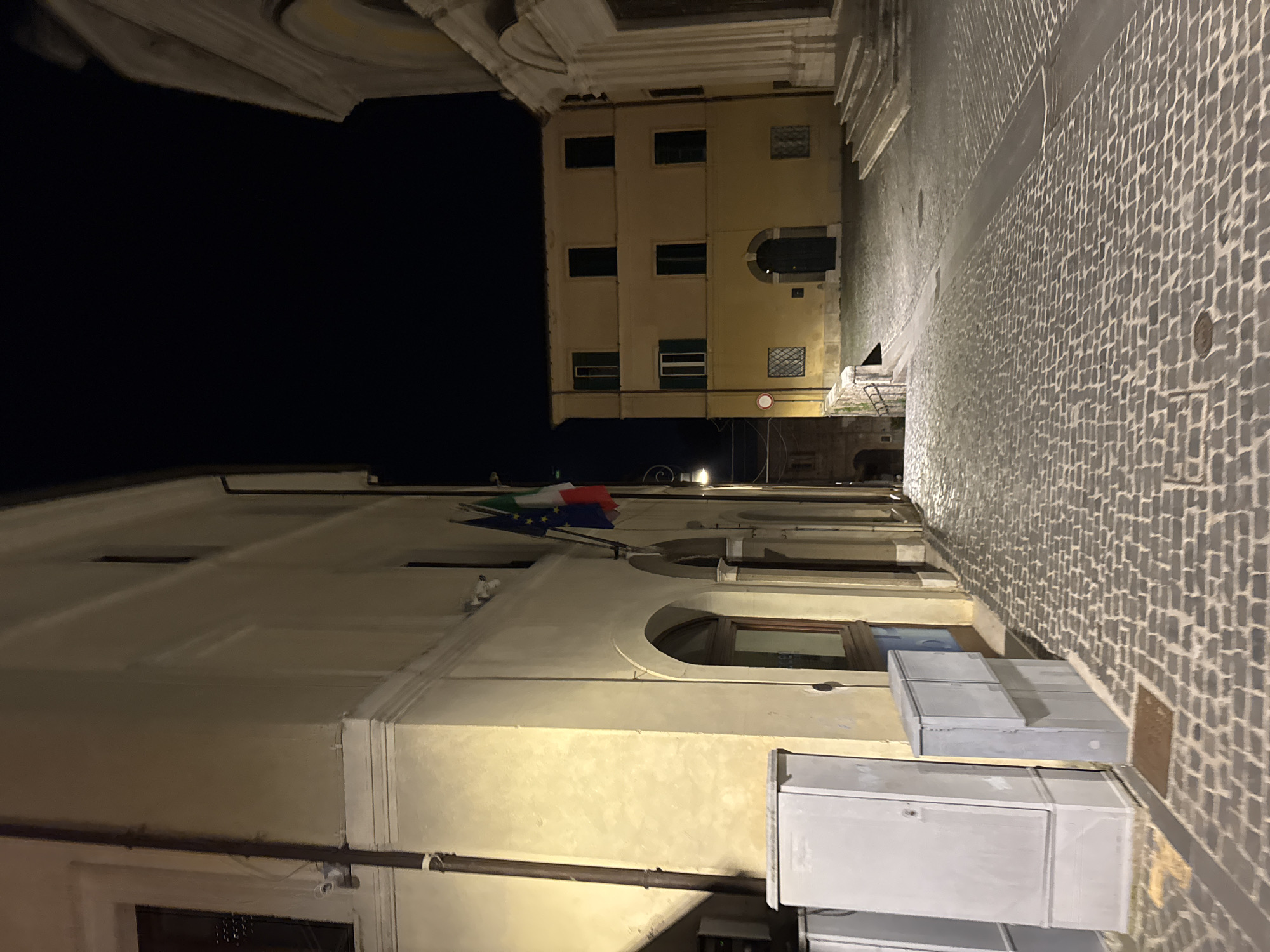 At night in Castelnuovo di Porto, Italy, a narrow cobblestone street runs between buildings. The building on the right is a pale yellow color with rows of windows. The building on the left is lighter in color and has a few windows and at least two white, box-like structures attached to its side. Flags, possibly those of Italy and the European Union, are affixed to the building on the left. A pipe runs along the wall of this building. The sky is dark.
